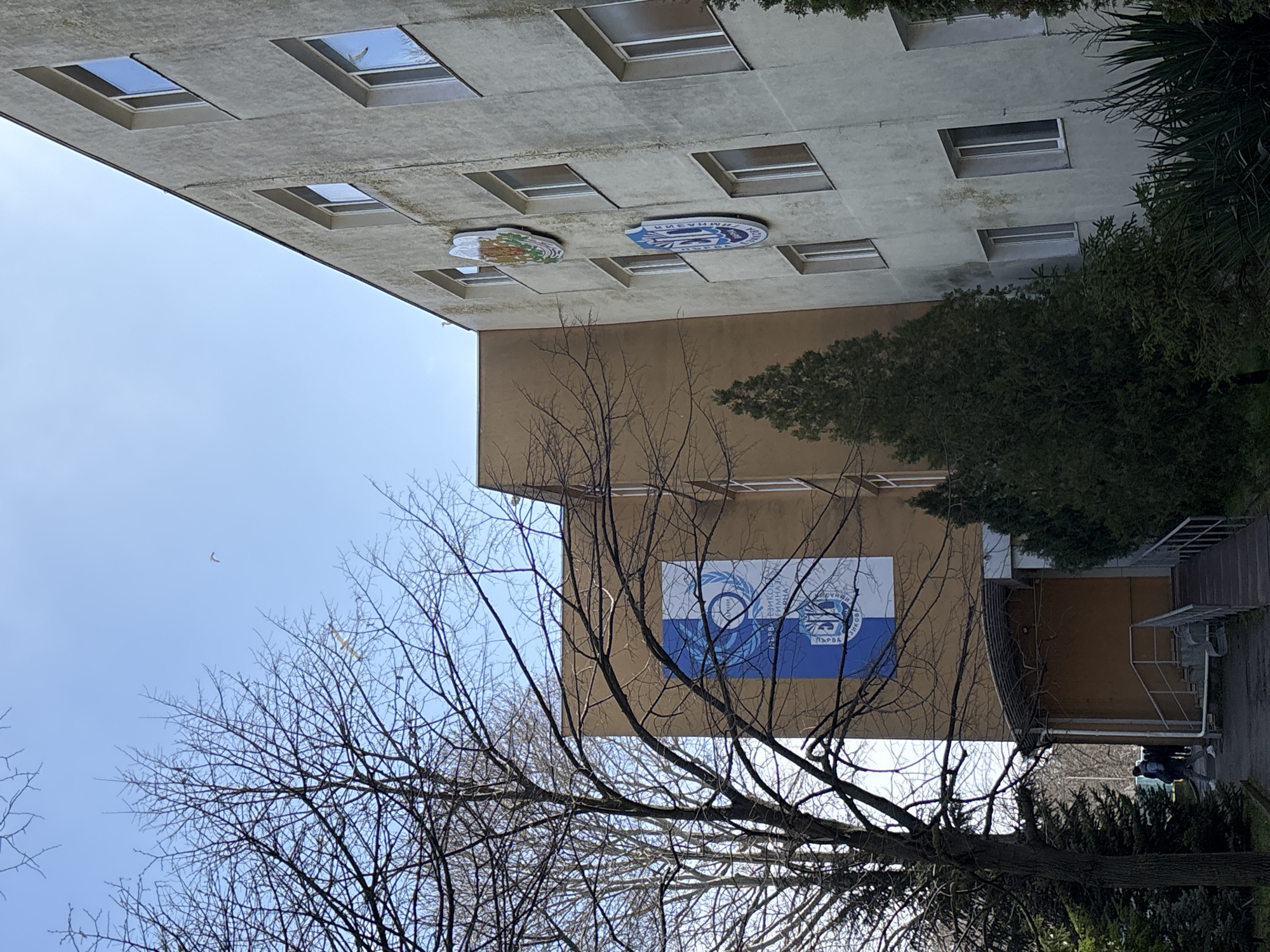 The image depicts a building in Varna, Bulgaria. The two-story building has a light gray upper level with a series of square windows, interspersed with two circular plaques of unknown insignia. The lower level is brown with a large blue and white banner featuring an image of a stylized eye. In the foreground, a leafless tree obscures the building and the sky. Some green foliage is visible on the right side of the image. A person wearing a dark jacket and white shoes is walking near the entrance of the building.