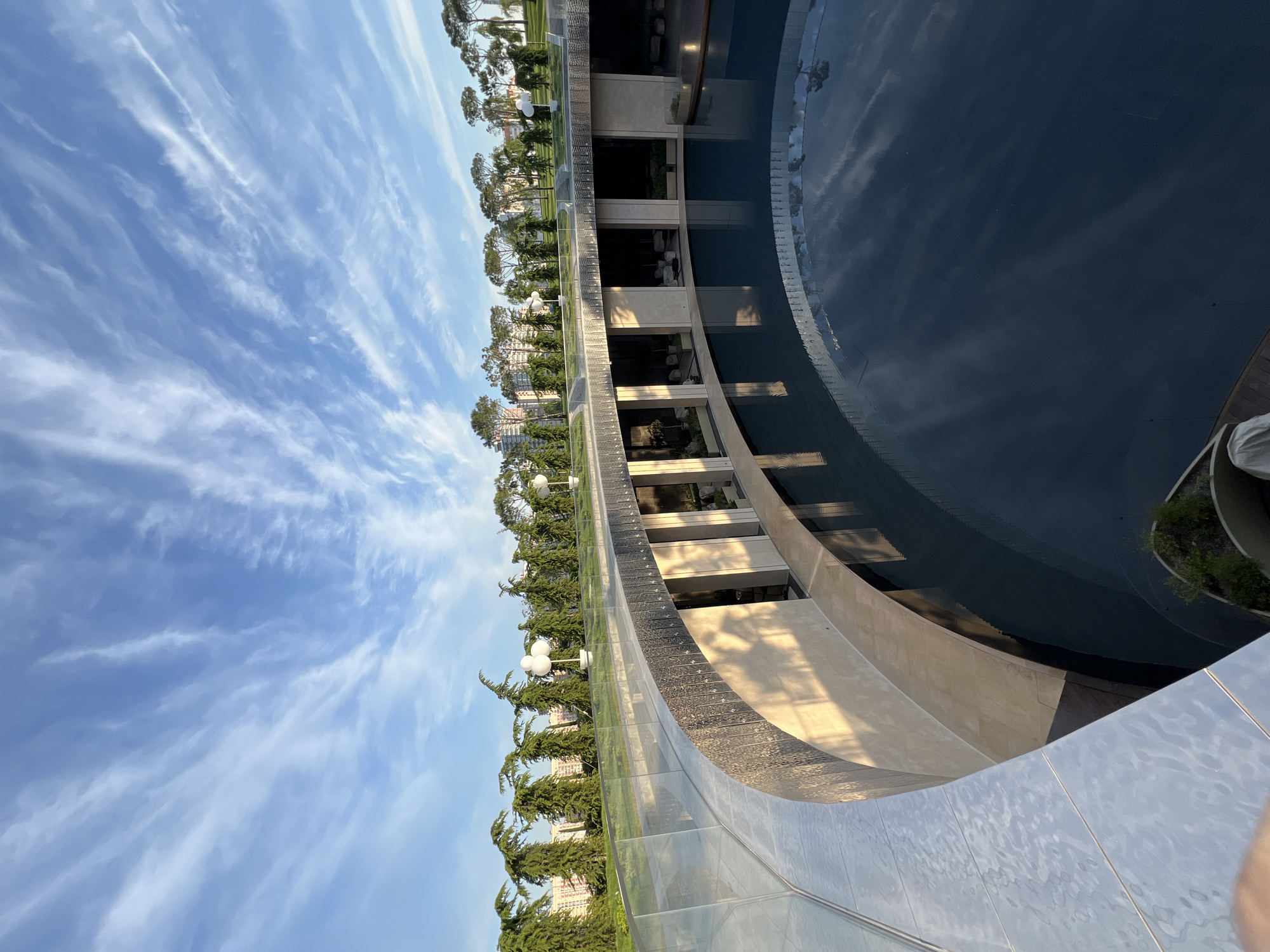The image shows a circular courtyard with a dark blue pool in the center, located in Krasnodar, Russia. The pool is surrounded by a curved walkway with an overhead glass railing. The walkway leads to a colonnade with arched openings that open into a shaded interior space. Lush green trees line the perimeter of the courtyard, with a few buildings visible in the background. The sky is blue with wispy white clouds, suggesting it is daytime. The scene appears to be a serene and modern outdoor recreational area.
