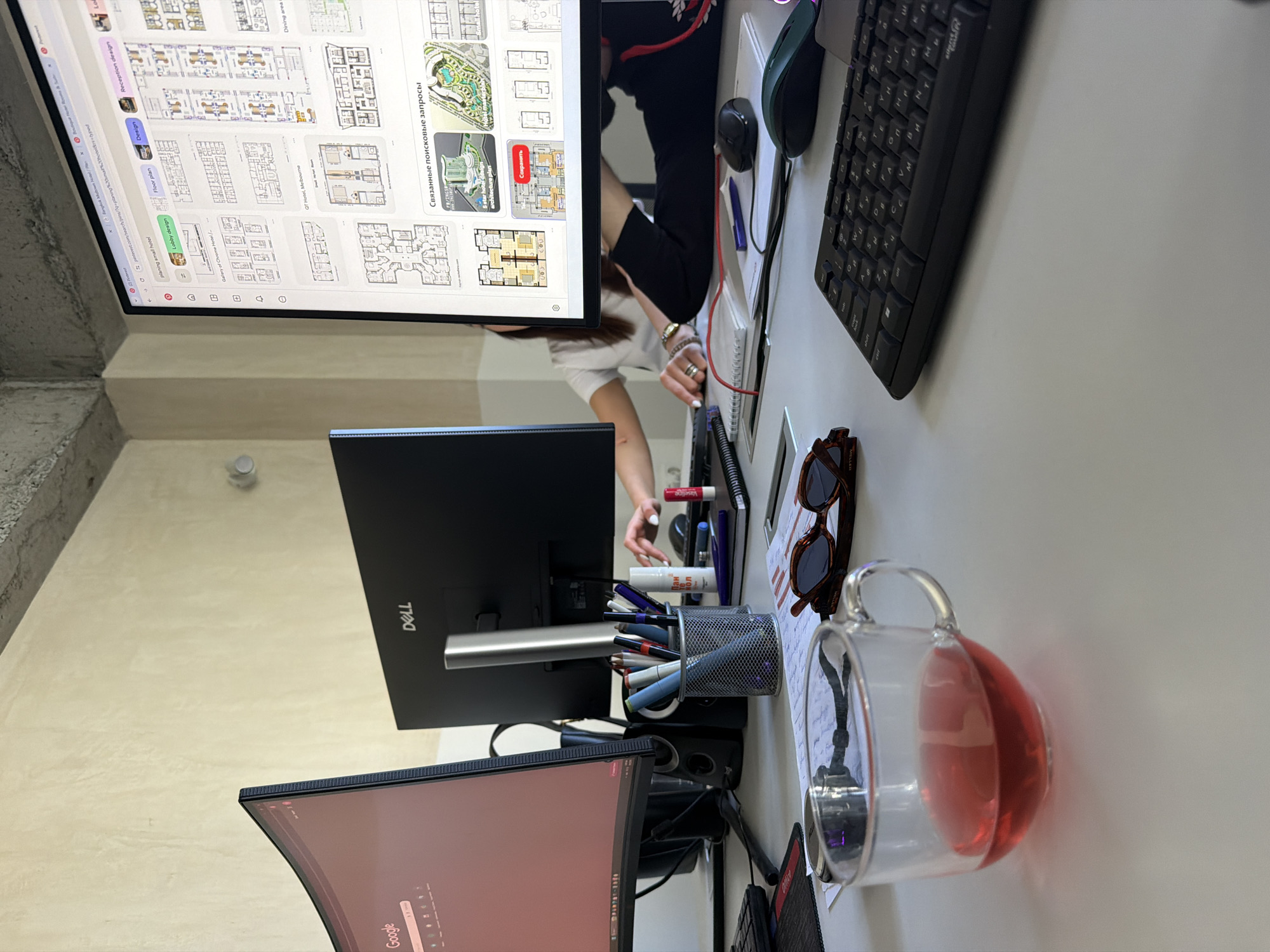 Here is a forensic description of the image:
The image shows a desk with multiple monitors. The top monitor displays a Pinterest page featuring floor plans. A person is sitting at the desk, interacting with the monitors. Various objects are present, including a keyboard, a mouse, sunglasses, a pen holder filled with pens and markers, a cup of tea or juice, and various other desk items. The environment appears to be a workspace with a concrete structural element visible in the background. There is no information in the image to identify the location in Yerevan, Armenia, or any other city.