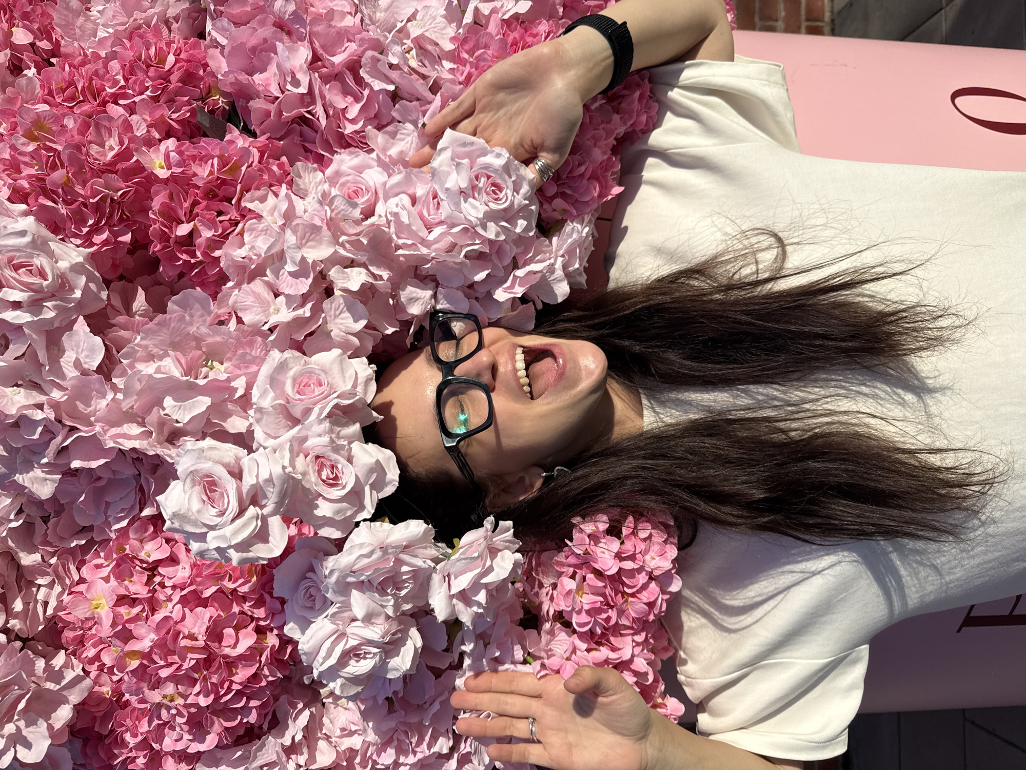 A woman with long dark hair and glasses smiles with her mouth open wide while lying down amidst a dense arrangement of pink and white artificial flowers. Her left hand is raised, palm facing forward, and her right hand is resting on the flowers near her face. She is wearing a white t-shirt. The background consists entirely of the flowers, which are a mix of rose and hydrangea-like blooms in various shades of pink and white. The image is cropped tightly, focusing on the woman and the floral backdrop. No specific location in Kazan, Russia is identifiable from the image.