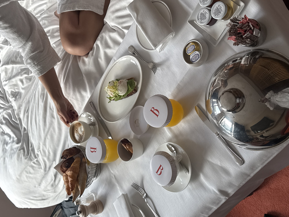 A breakfast scene unfolds on a white tablecloth covering a tray situated on a bed. A person, partially visible in the upper right, wears a white terry cloth robe, with their bare legs also visible. Their right hand reaches towards a white ceramic cup containing a beverage with foam, likely coffee or cappuccino, on a matching saucer.
The tray holds an array of breakfast items and serving ware. In the upper left, a silver-colored wire basket contains several pastries, including croissants and baguette-like bread. Adjacent to this is a wooden pepper mill and salt shaker. Below them, a white folded napkin and a fork are positioned next to an empty plate.
Further right, a small glass jar with a white lid bearing a red "B" logo holds orange juice. Next to it, a small white bowl contains what appear to be sausages. An oval white plate with a dark rim holds a piece of dark toast topped with avocado spread, two poached eggs, and greens. A fork is placed to the right of this plate.
Towards the bottom center, another white ceramic cup with a similar "B" logoed lid sits on a saucer with a spoon. Another orange juice jar with a "B" logo is placed nearby. A silver-colored knife lies next to a large, domed silver-colored serving cover, which reflects the photographer, clad in a white robe, holding a phone. To the right of the serving cover, a small white bowl contains two individually wrapped butter portions. A rectangular white dish holds three small glass jars of what appear to be jams or spreads, and another small container holds an assortment of sugar packets and stirrers. The bed features rumpled white linens. A reddish-orange carpet or rug is partially visible on the far left.
