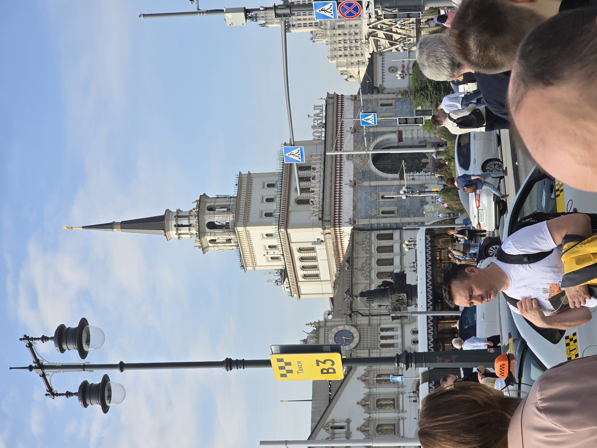 The image depicts a street scene in Moscow, Russia, with the Kazansky railway station prominently displayed. The station features a distinct clock tower and ornate facade. Several people are present, including one man wearing a white t-shirt. Taxis are also visible, along with a taxi stand sign displaying "Taxi B3". The sky is blue with light clouds. Additional buildings can be seen in the background.
