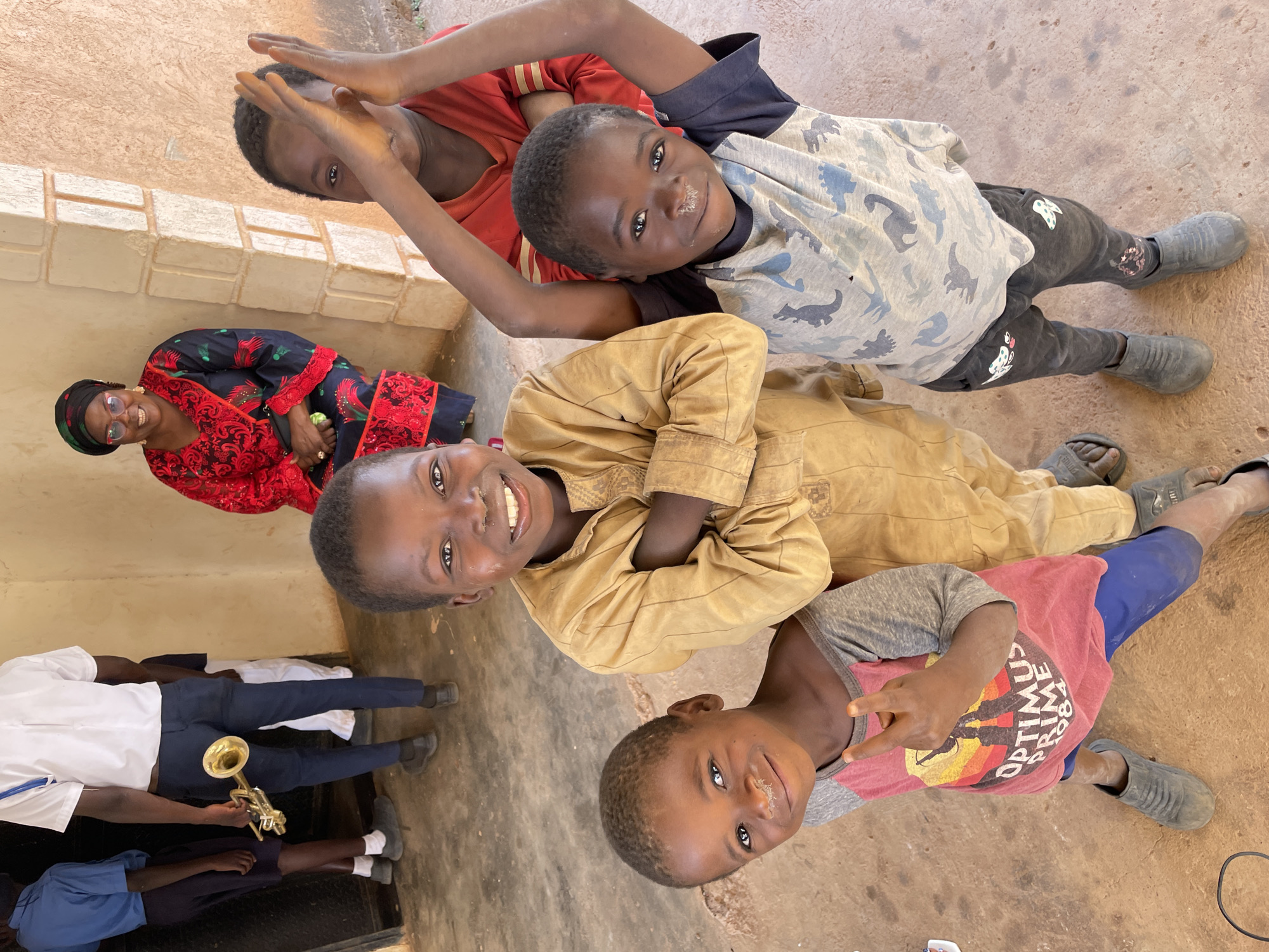 This overhead shot captures a joyful group of people, primarily children, looking up at the camera in what appears to be an indoor or sheltered outdoor setting in Ladura Foron, Nigeria. The ground is a light brown, possibly concrete or hard-packed dirt, and the background features light-colored, rough-textured walls, suggesting a modest building.
At the top, partially visible, a child in a red shirt with hands raised near their face looks directly at the camera with a smile. Below them, another young boy wears a light grey t-shirt with blue dinosaur patterns and dark grey trousers adorned with white, perhaps cartoon character, motifs. He smiles broadly. Next to him, a boy in a light golden-brown, traditional long-sleeved tunic (likely a kaftan) also smiles, revealing his teeth.
Further down, a boy in a grey t-shirt with a colorful graphic print, blue shorts, and dark clogs flashes a peace sign with one hand. The graphic on his shirt clearly reads "OPTIMUS PRIME 1984".
An adult woman is visible on the left, wearing a black and red patterned dress with a matching headscarf and glasses. She is smiling warmly. Behind her, partially obscured, is an adult male in a white shirt and dark trousers, holding a brass musical instrument that appears to be a trumpet. Another child in a blue uniform top is also partially seen near the adult with the trumpet.
The overall mood is cheerful, with many subjects displaying bright smiles and engaging directly with the camera. The natural, bright lighting indicates it is daytime. The presence of a musical instrument hints at a possible performance or practice session, or simply the daily life of people associated with music.