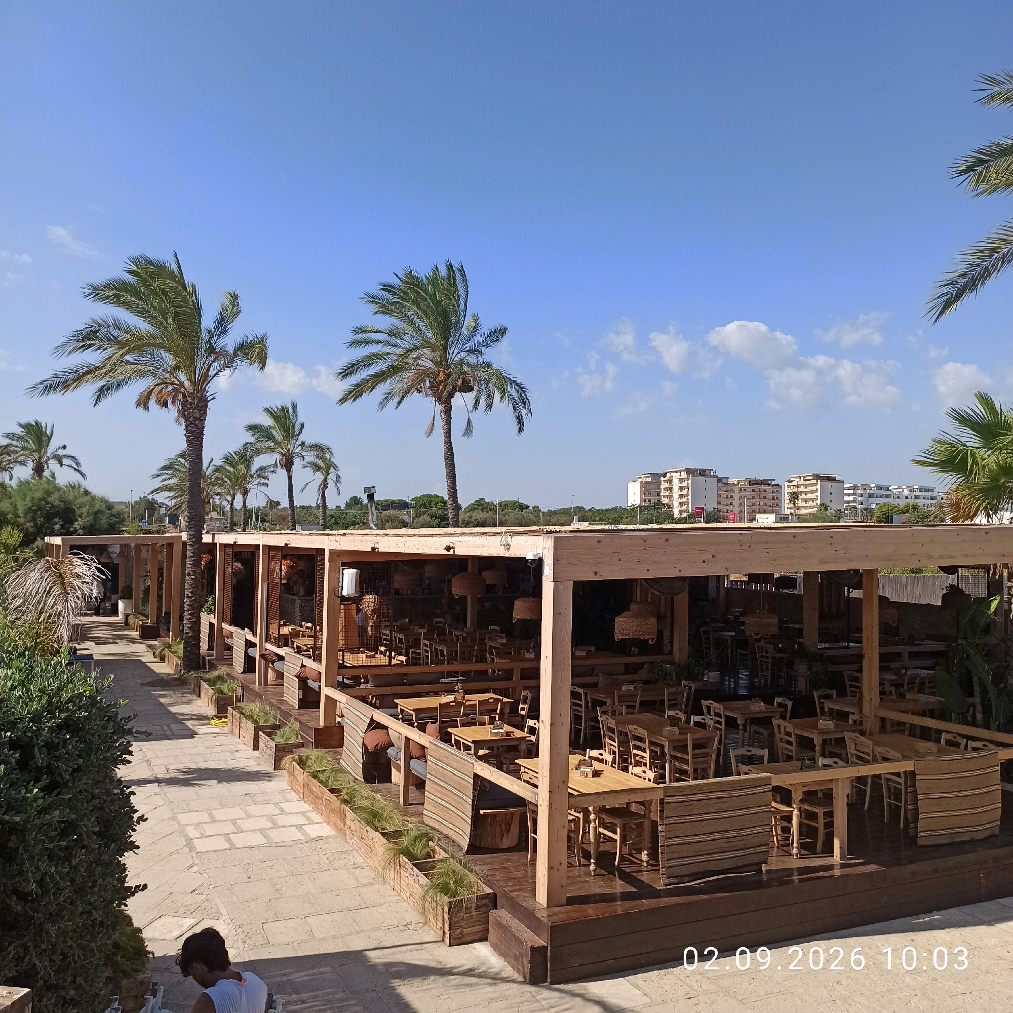 The image depicts an outdoor dining area of a restaurant located in Molfetta, Italy. Palm trees are visible throughout the scene, indicating a Mediterranean climate. The restaurant features a wooden structure with an open-air seating arrangement, furnished with numerous wooden tables and chairs. Decorative elements such as woven lampshades and striped fabric panels are present. The ground is paved with stone tiles. In the background, several apartment buildings are visible, suggesting an urban or semi-urban setting adjacent to the dining establishment. A date and timestamp, "02.09.2026 10:03," is superimposed on the lower right corner of the image. One person, a young male wearing a white t-shirt, is partially visible in the foreground on the left, appearing to be seated or stooping.