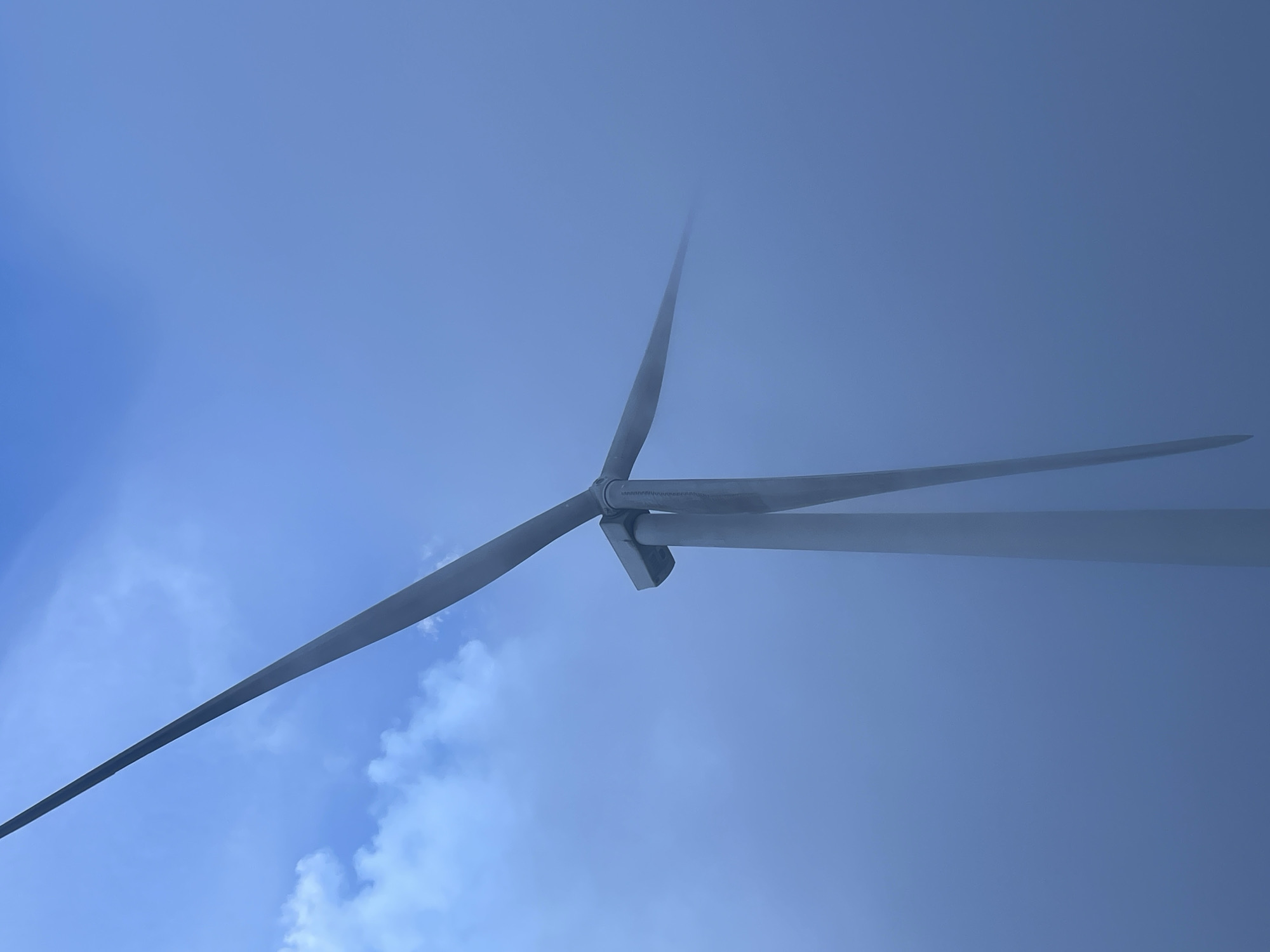 A low-angle shot of a wind turbine with three blades against a blue sky partially obscured by fog. The turbine appears to be located in an open area, as indicated by the expansive sky. The image quality suggests daylight conditions, though the fog may dim the overall illumination. There is no evidence of people or specific objects identifying the precise location in Trường Thọ, Vietnam.