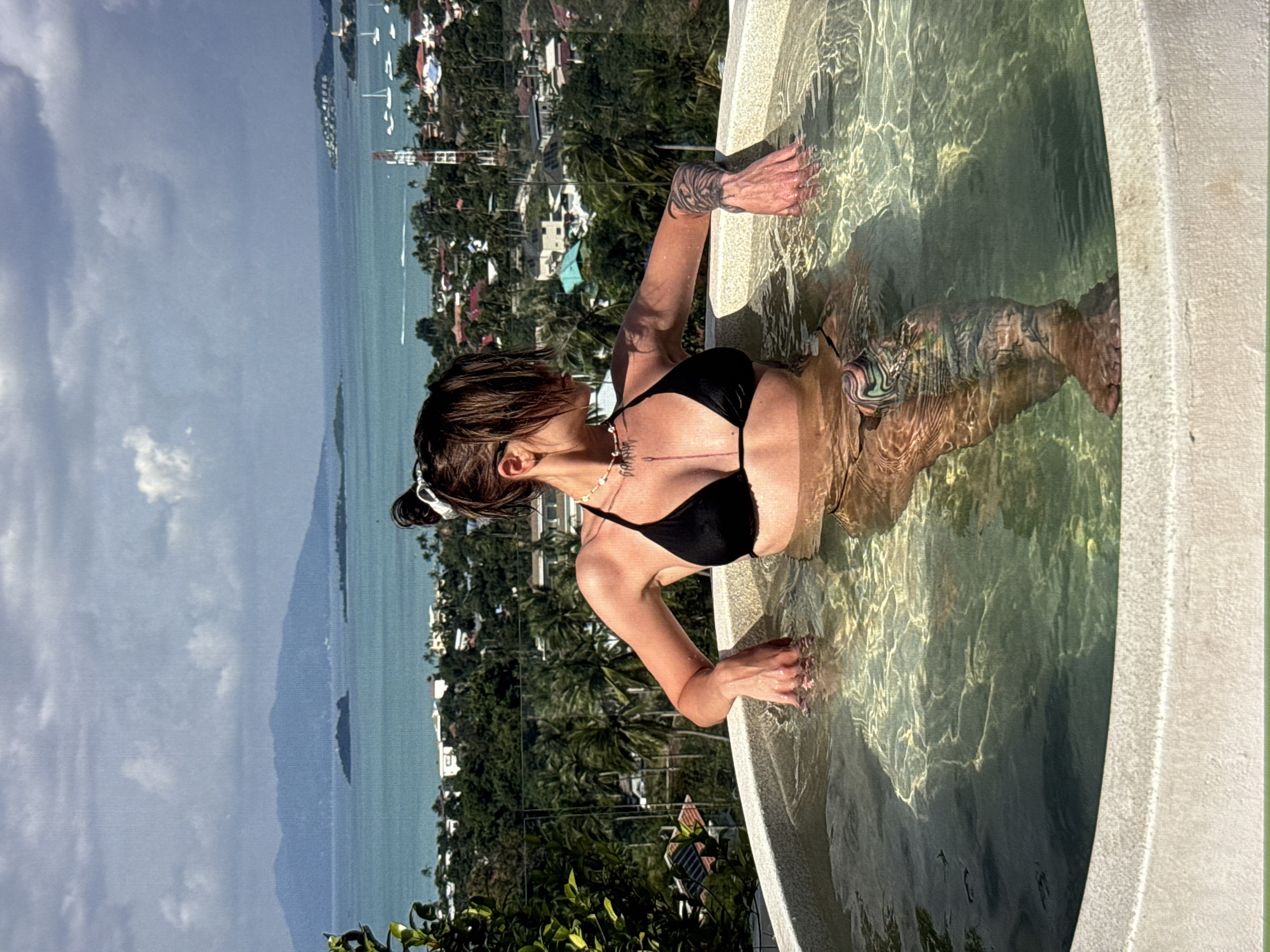 A young woman with dark hair in a black bikini is seated in a circular hot tub with clear water. The hot tub is located outdoors, with a panoramic view of a tropical landscape. In the background, there are lush green trees, buildings, and a body of water with islands and boats. The sky is partly cloudy. The woman is looking away from the camera, towards the view. She has tattoos on her arms and legs. The scene is brightly lit by natural sunlight. The location is not Moscow, Russia; it is a tropical environment.
