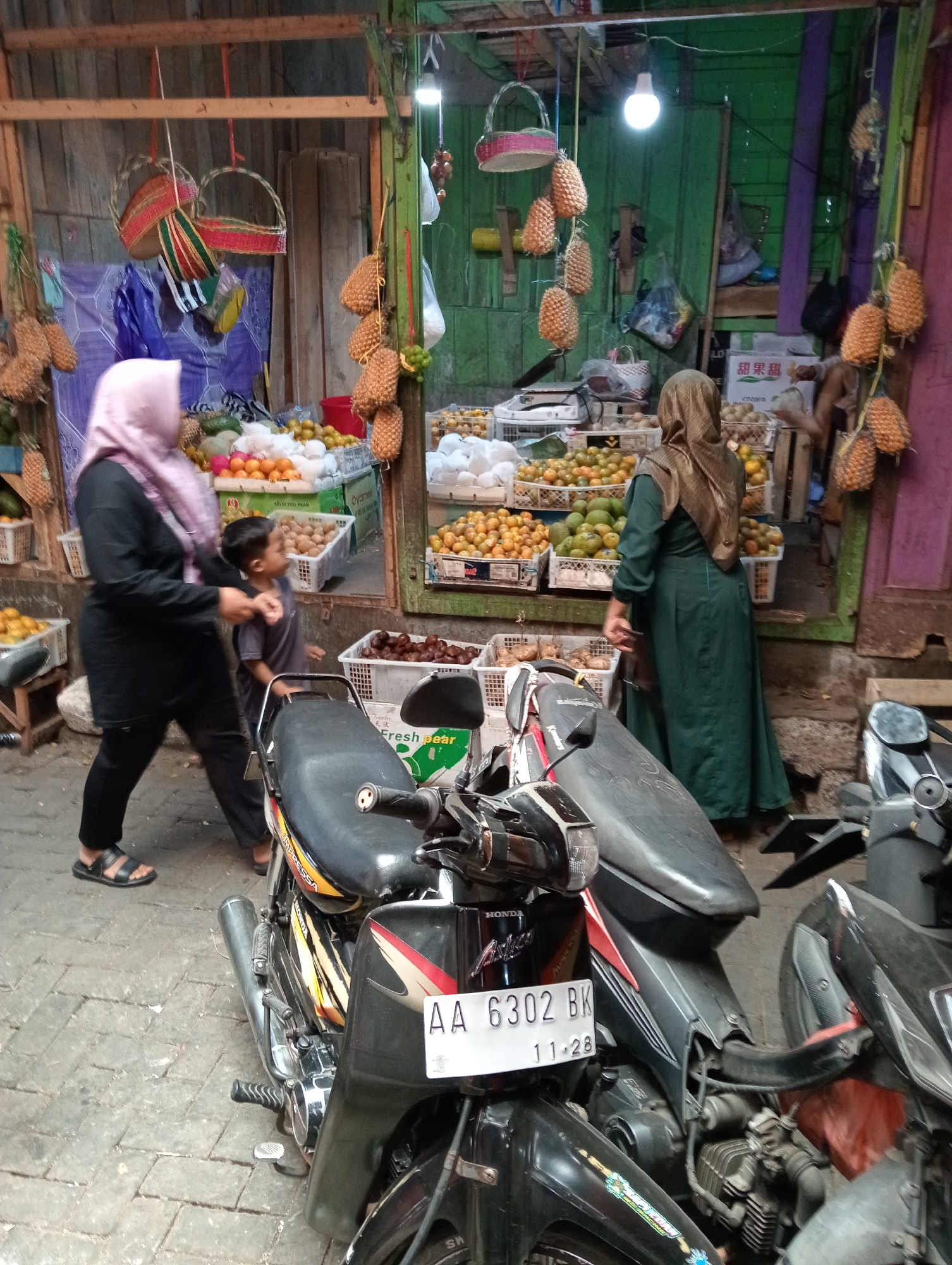 This image depicts a street-side fruit stand in Grabag, Indonesia, bustling with activity and goods. Two women, both wearing hijabs, are present. One woman, wearing a pink hijab and black clothing, is walking away from the camera. The other woman, in a dark green dress and a tan hijab, is facing away from the camera, appearing to be interacting with the fruit stand. A young boy in a gray t-shirt is walking next to the woman in pink.
The fruit stand is laden with various produce, including oranges, mangoes, and what appear to be pineapples hanging from the ceiling. Baskets and crates are used to display the fruits. Several motorcycles are parked in front of the stand, with one prominently in the foreground displaying a license plate with the characters "AA 6302 BK" and "11.28". The environment suggests a local marketplace setting, with wooden structures and a rough, paved ground. The overall atmosphere is that of a vibrant, everyday scene in a Indonesian city.