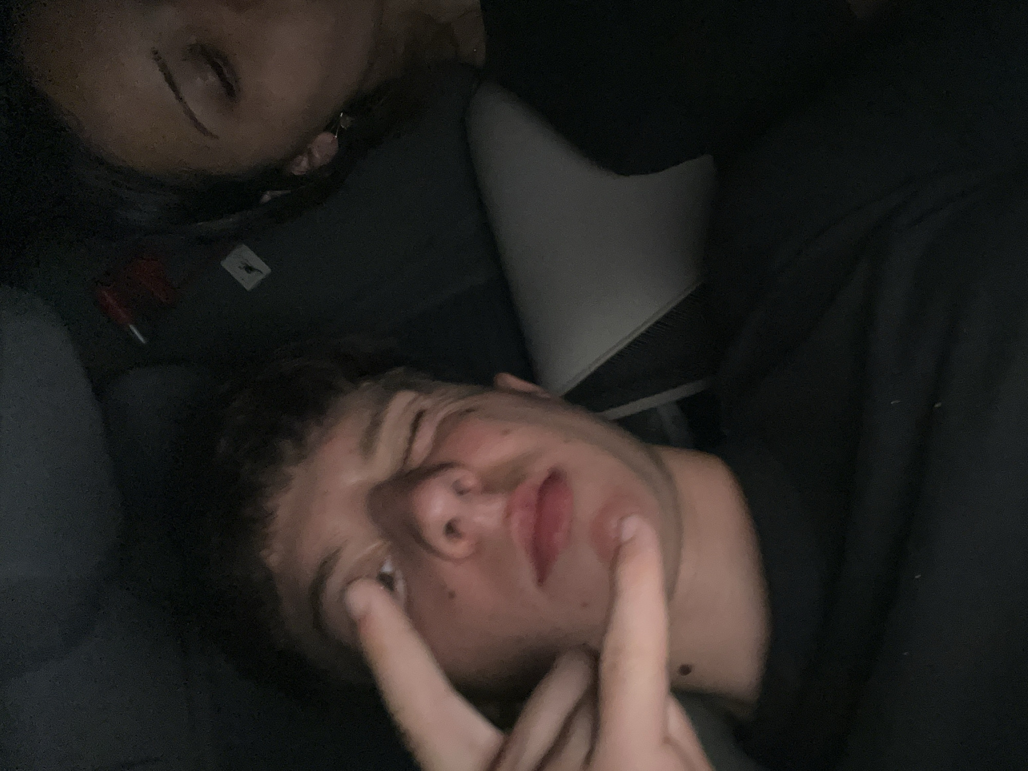 Two individuals are present in an indoor setting. One is lying prone and appears to be making a peace sign with their hand. The other individual is above them and appears to be asleep. The location is not discernible from the image.