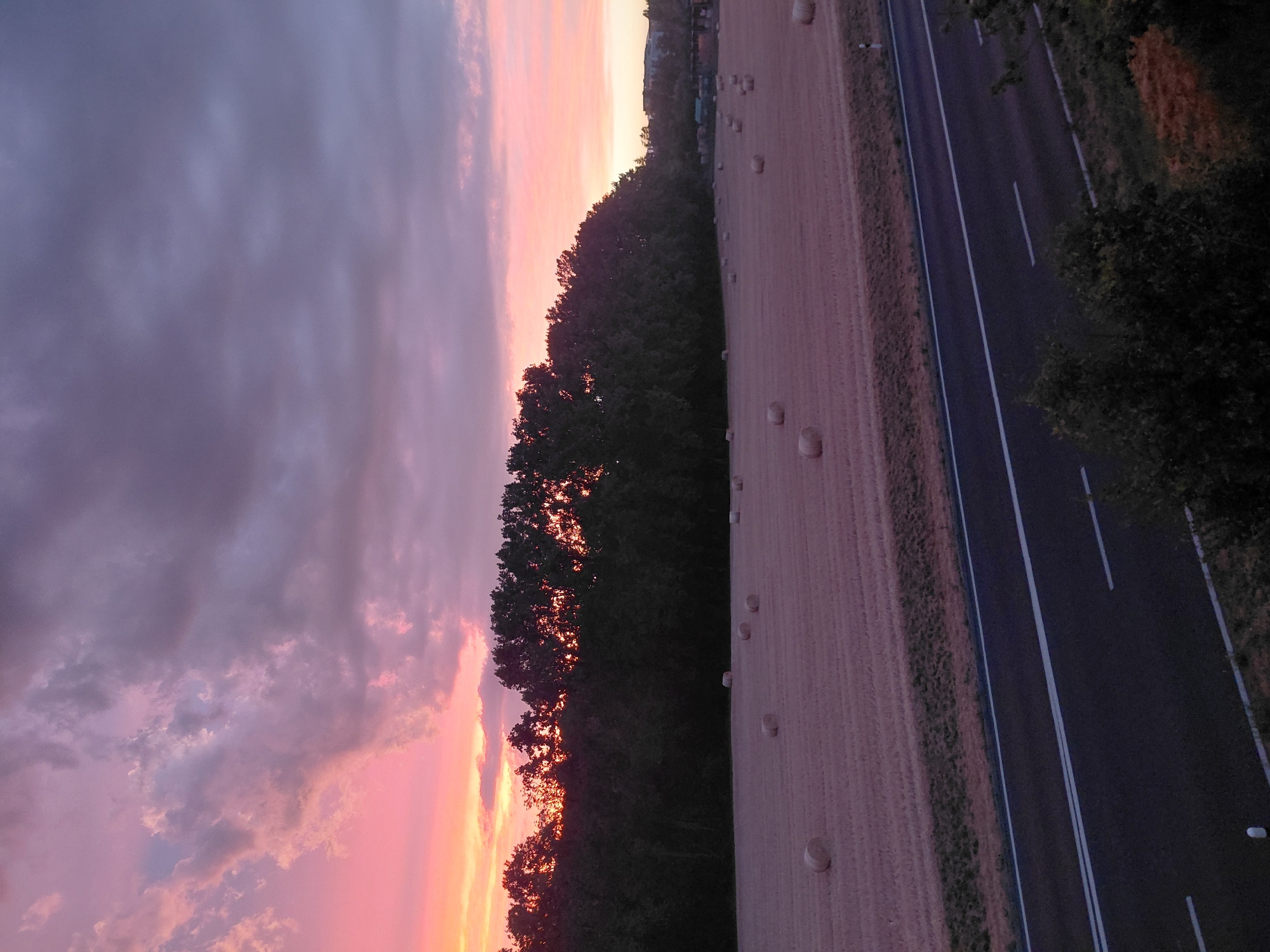 The image depicts a rural landscape at dusk, featuring a road in the foreground, a field of hay bales, and a line of trees against a dramatic sky. The sky is filled with clouds, colored in hues of pink, purple, and gray, indicating the sun has recently set or is about to set. The field contains numerous round hay bales scattered across it, arranged in an irregular pattern. A dark asphalt road with white lane markings runs horizontally across the lower portion of the image. Behind the field, a dense line of trees forms a dark silhouette against the colorful sky. In the distance, buildings are faintly visible, suggesting the proximity of a settlement. There are no people or animals visible in the scene. The overall impression is one of tranquility and natural beauty.