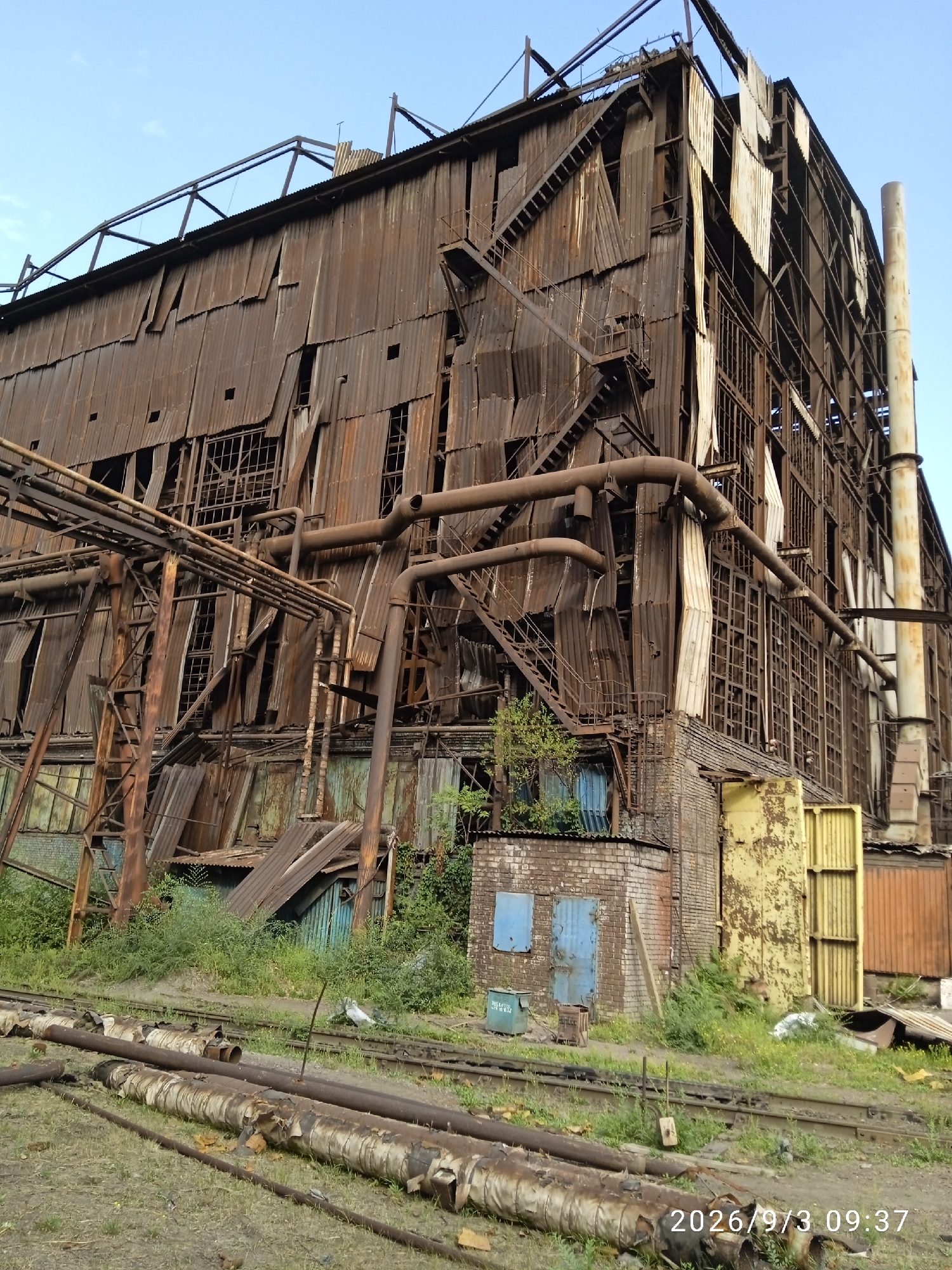 The image shows a large, dilapidated industrial building with extensive rust and damage to its corrugated metal siding. The building has multiple levels, exposed structural elements, and numerous broken windows. Several large pipes are visible running along the exterior of the structure, some appearing to be part of an industrial process. Outside the building, there are railway tracks and a pile of discarded pipes covered in peeling material. Overgrown vegetation is present around the base of the building and between the tracks. The sky is clear and blue. The timestamp "2026/9/3 09:37" is overlaid on the bottom right corner of the image.