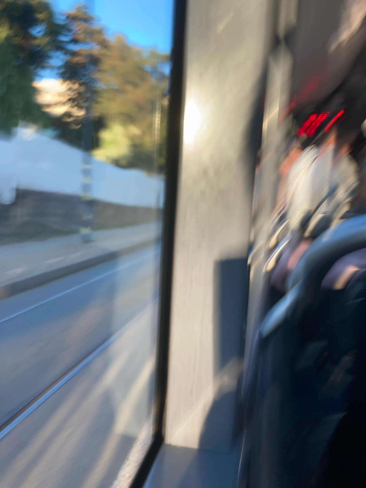 The image shows an interior view of a moving vehicle, likely a tram or bus, looking out of a window. The perspective is from within the vehicle, with a window on the left showing a blurred outdoor scene of trees, a road, and a sidewalk under a blue sky. The right side of the image is dominated by the interior of the vehicle, with blurred seats and what appear to be red digital displays or lights in the upper right quadrant. The lighting suggests it is daytime, possibly late afternoon due to the warm tones in the outdoor scene. The overall image is characterized by motion blur, indicating the vehicle is in motion. Without any specific landmarks or identifying features, it is not possible to definitively place this scene in Prague, Czechia.