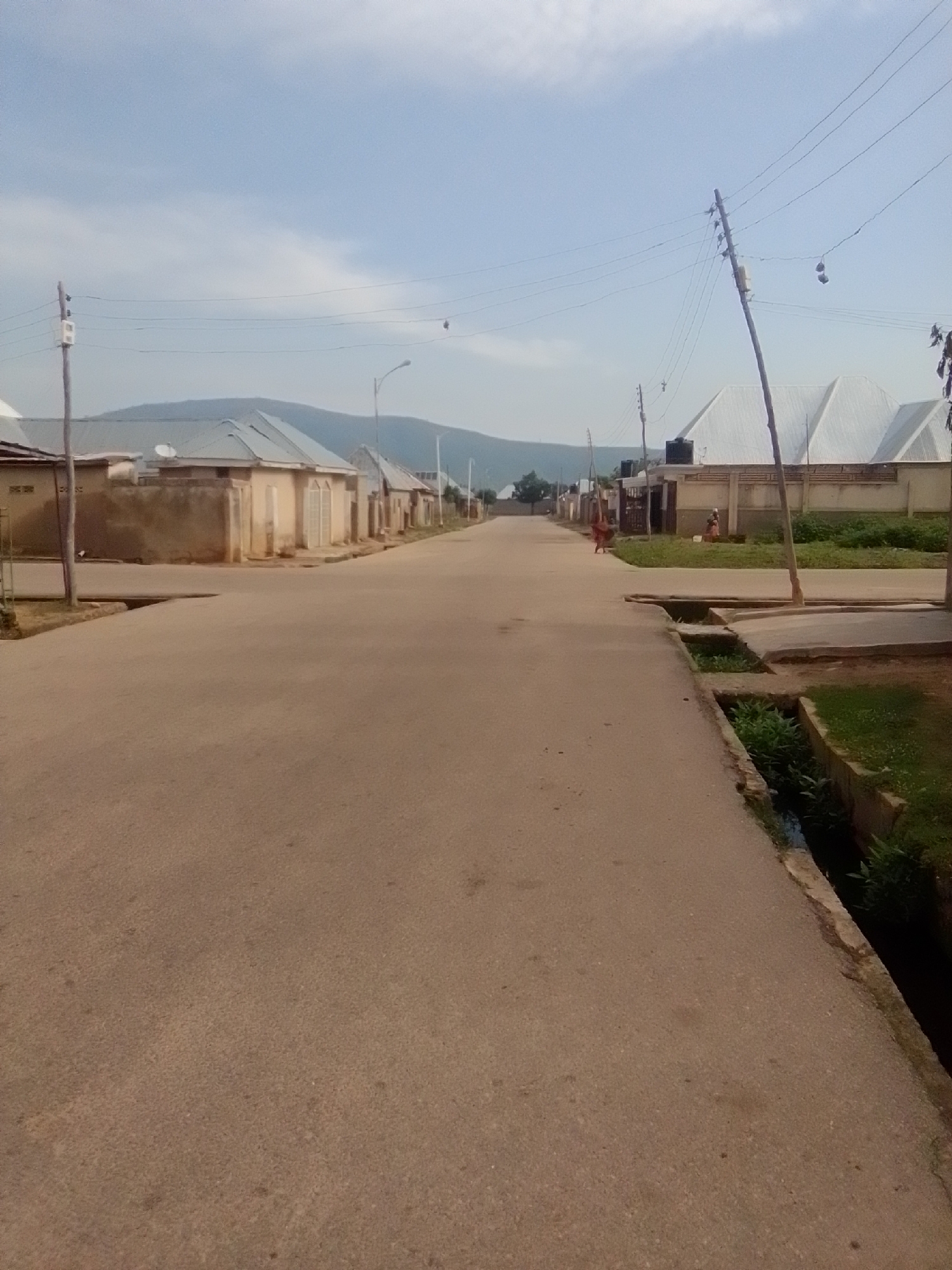 The image depicts a street scene in what appears to be a residential area. The street is paved and appears to be relatively clean. On either side of the street are houses with light-colored walls and tiled roofs. Some houses have fences or walls surrounding them. There are power lines and utility poles visible, suggesting access to electricity. In the distance, a range of hills or mountains is visible under a partly cloudy sky. There are a few people visible in the image, including one person walking on the street and another standing near a house. A concrete drainage ditch runs alongside the road in some sections, with some vegetation growing in it. The overall impression is one of a quiet, functional neighborhood.