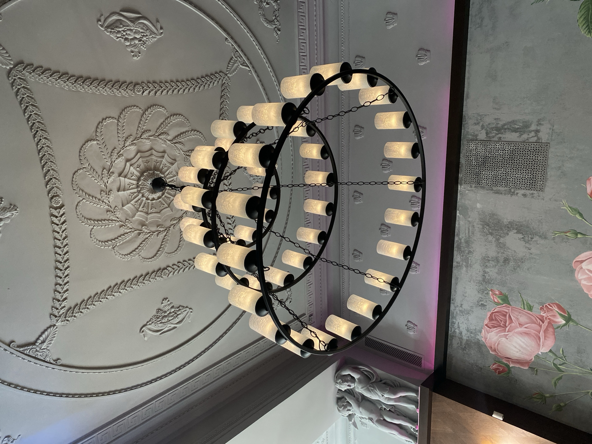 Here is a description of the image:
The image depicts a ceiling view, showcasing a decorative ceiling with ornate plasterwork details. A prominent chandelier with a circular design and multiple cylindrical lights hangs from the center of the decorative element. The color scheme is primarily white and gray, with accents of light from the chandelier and some colorful wall decorations. The wall has rose motifs and an ornate white sculpture. A vent is also visible.