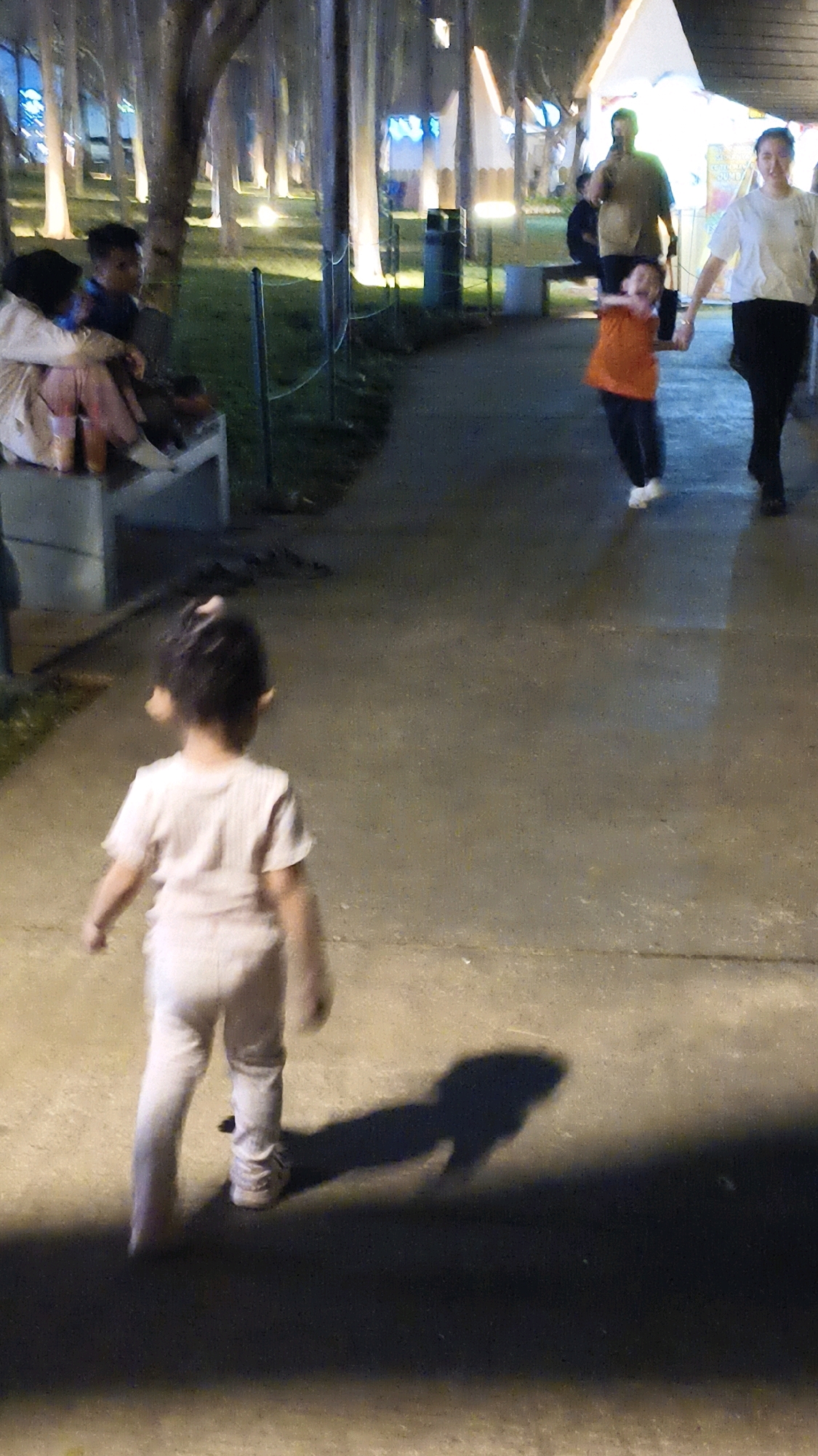 The image depicts a nighttime scene in a park-like setting, likely in the city of West Jakarta, Indonesia, given the presence of tropical trees and the general ambiance. The foreground features a young child, viewed from behind, walking away from the camera on a paved walkway. The child is wearing a light-colored t-shirt and pants. A long shadow of the child is cast on the ground. Further in the background, to the left, a couple is seated on a bench, with one person leaning against the other. They appear to be wearing casual attire. In the mid-ground and background, several other individuals are visible. To the right, a woman in a white t-shirt and black pants is walking, holding the hand of a young child in an orange shirt. Behind them, a man in a light-colored shirt is walking. The environment is illuminated by artificial lights, possibly from streetlights or temporary fixtures, which cast a warm glow on the trees and ground, creating a soft, diffused lighting effect. Several large trees are prominent in the background, lining the walkway. Tents or temporary structures are visible in the far background, suggesting a possible event or market. The overall impression is a relaxed evening outing in a public space.