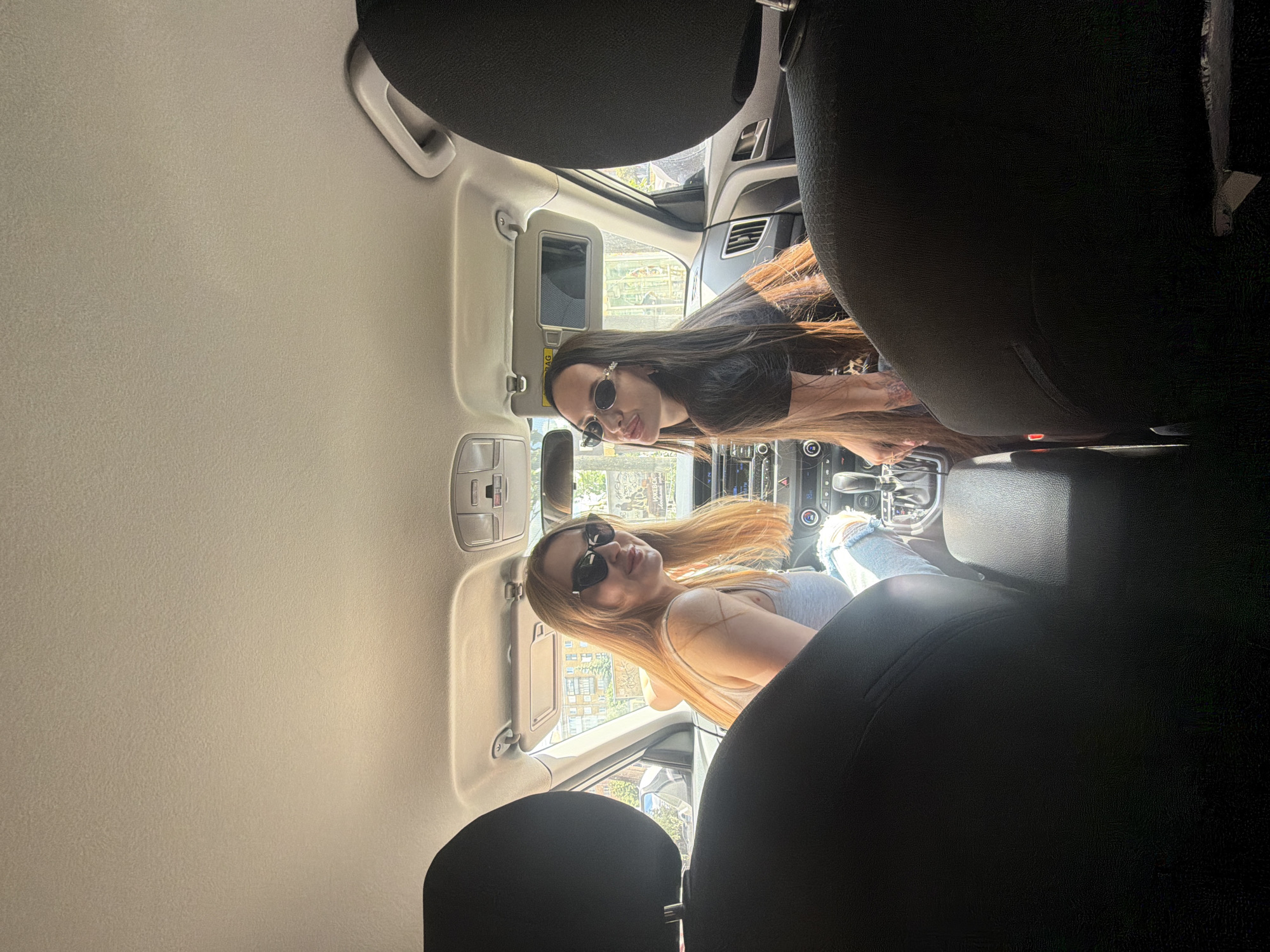 The media file presents an interior view of a car, captured from the perspective of the back seat looking forward. Two young women are the primary subjects, seated in the front passenger and driver's seats, respectively.
The woman in the passenger seat has long, straight dark brown hair and wears dark sunglasses, a dark short-sleeved top, and an earring on her left ear. She is turned towards the camera, leaning slightly forward, with a subtle smile.
In the driver's seat, a woman with long, blonde or light brown hair is visible. She also wears dark sunglasses and a light-colored tank top. Her leg, dressed in light blue distressed jeans, is visible near the central console. Like her companion, she is turned towards the camera and displays a gentle smile.
The car's interior features a light-colored headliner and overhead console. A sun visor on the passenger side is flipped down, showing a mirror, and the word "AIRBAG" is faintly visible on it. Parts of the dashboard, air vents, and the central console with an automatic gear shifter are also visible. The headrests of the front seats are in the foreground, framing the view of the women.
Outside the car windows, bright daylight illuminates the scene, indicating it is daytime. Elements of an urban environment, including buildings and some indistinct graffiti or text on an external structure, are visible. The overall atmosphere conveyed is relaxed and pleasant, with both women appearing cheerful. The setting for this image is Izhevsk, Russia.