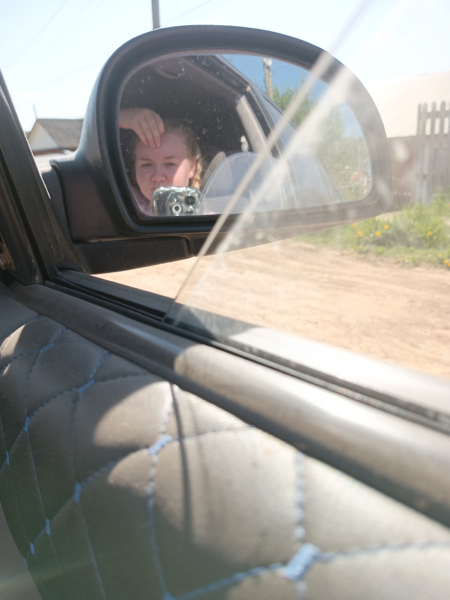 The image shows a selfie reflected in a car's side mirror. The person in the mirror is holding a smartphone and touching their forehead. The car is driving along a dirt road, with a wooden fence and greenery visible in the reflection. A house with a grey roof can also be seen in the background. The interior of the car features quilted door panels with light blue stitching. The location is likely Фоки, Russia, based on the prompt.