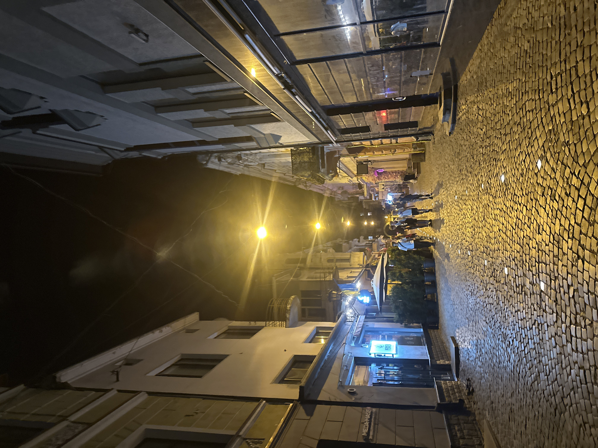 The image depicts a cobblestone street at night in Ternopil, Ukraine. The street is lined with buildings on both sides, appearing to be a commercial area with shops and possibly restaurants. Bright yellow streetlights illuminate the scene, casting long shadows on the textured ground. Several people are walking down the street, silhouetted against the light. Some of the buildings have visible signage, and one building on the left has a sign that appears to read "MEGA-MASS". There are also umbrellas and potted plants visible outside some establishments, suggesting outdoor seating areas. The overall atmosphere is that of a lively urban street after dark.