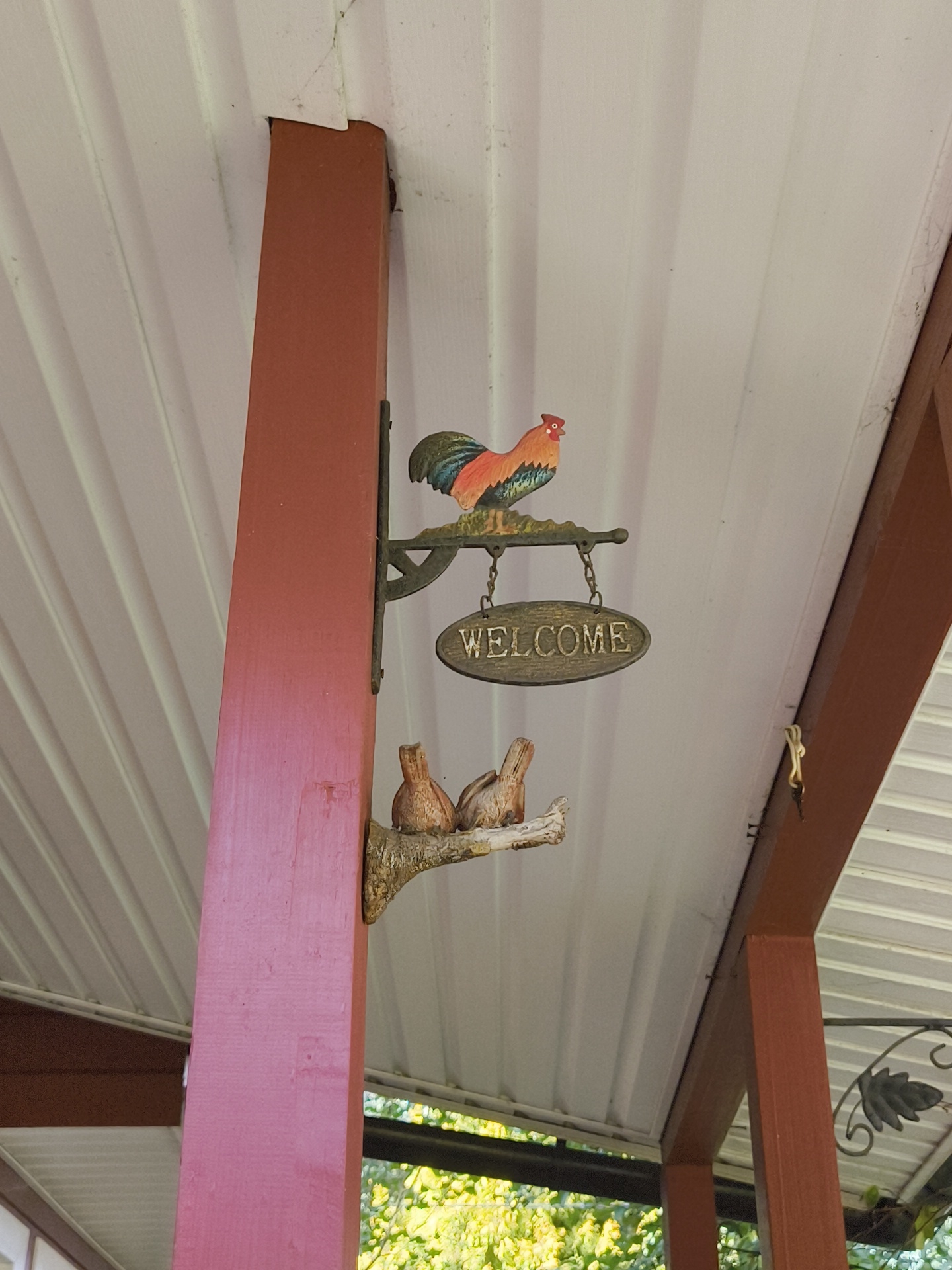 The image shows a porch with a red-painted post supporting the white corrugated roof. Attached to the post is a decorative metal sign featuring a rooster at the top and the word "WELCOME" on an oval plaque below. Further down the post, a decorative branch with two small bird figures is mounted. In the background, lush green trees are visible, indicating an outdoor setting. The scene is devoid of people and the overall impression is one of a welcoming, rustic porch decoration. The location is identified as Правдист-1 Снт, Russia.