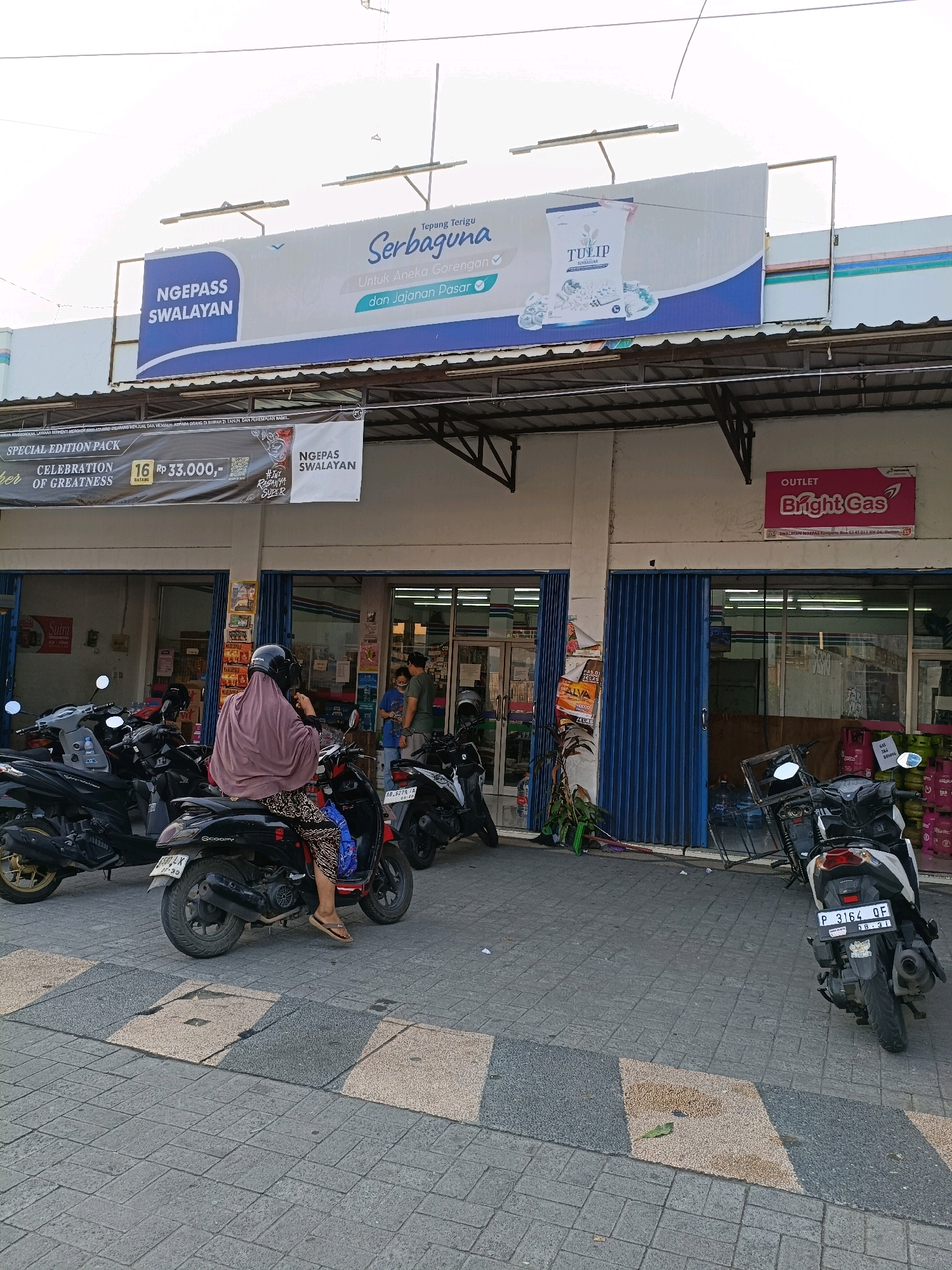 The image depicts the exterior of a convenience store, "NGEPASS SWALAYAN", located in Purwomartani, Indonesia. A large banner above the store advertises "Tepung Terigu Serbaguna Tulip", a type of all-purpose flour. In front of the store, several motorcycles are parked, and a person wearing a headscarf and patterned pants is seated on a red scooter. Two men are standing near the entrance of the store, one appears to be speaking with the other. The store has blue accordion-style doors and a sign indicating "OUTLET Bright Gas". The ground is paved with interlocking bricks. The lighting suggests it is daytime, possibly late afternoon.