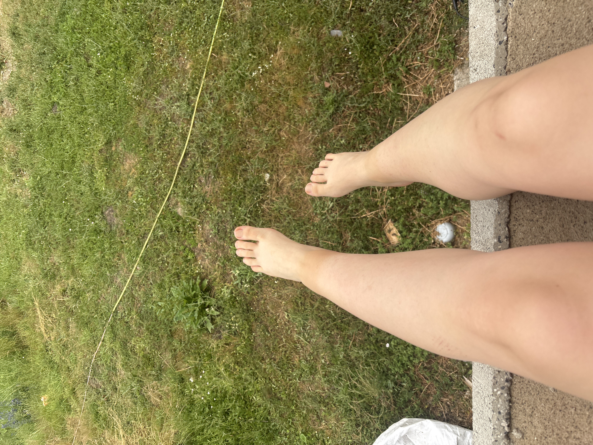 The media file features a close-up view of a person's legs and feet, positioned outdoors. The foreground is dominated by the person's fair-skinned legs and bare feet, which are angled towards the right side of the frame. The skin on the legs appears smooth and has a slight tan line or variation in coloration on the right leg. There are some faint marks or blemishes visible on the right thigh.
The legs and feet are situated on the edge of a paved surface, possibly a concrete walkway or patio, which is textured and speckled with gray and white aggregate. To the left of the paved surface is a grassy area, a lawn. The grass is green and appears somewhat uneven, with patches of drier, browner grass and some small white wildflowers interspersed. A thin, yellow string or cord runs diagonally across the grassy area from the upper left to the lower right. In the background of the grassy area, there are some dry leaves and twigs. A small white object, possibly a golf ball or similar item, is visible on the grass near the paved edge. A white, crinkled material, possibly a tarp or bag, is partially visible at the bottom right corner of the image.
The lighting suggests it is daytime, likely outdoors with natural light. The overall impression is that of a casual moment, perhaps someone resting outdoors. No specific activity is definitively depicted, other than the person's legs being positioned in the scene. There is no visible text in the media file. The setting appears to be a backyard or garden area.
