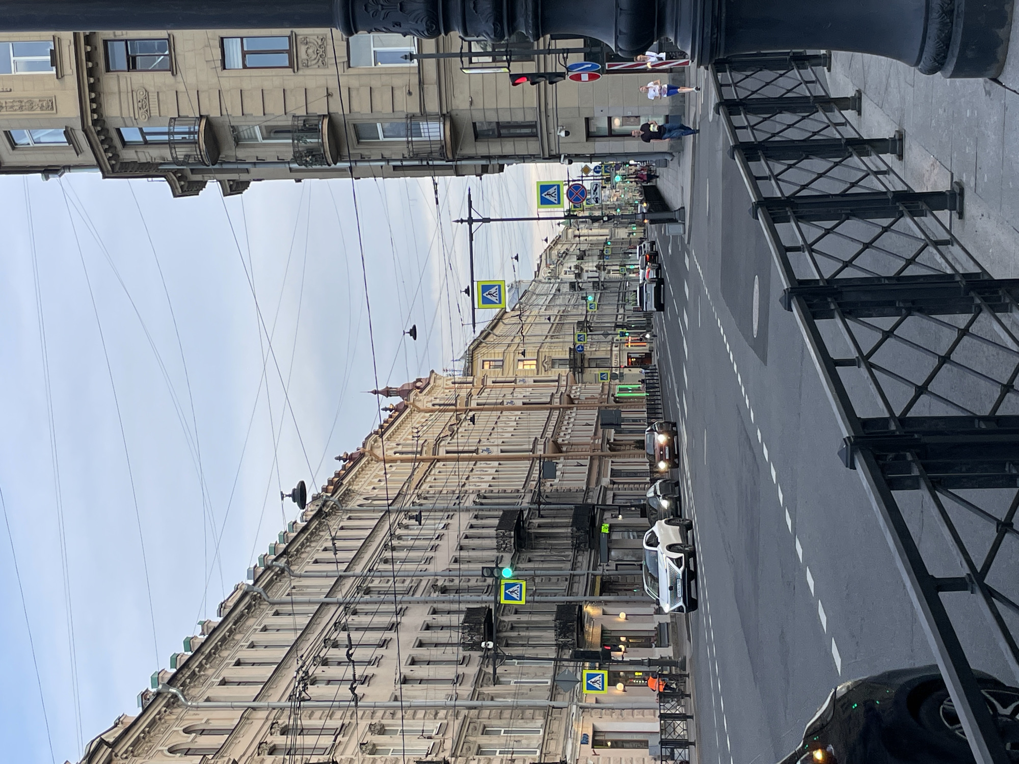 This image depicts a wide urban street in Saint Petersburg, Russia, observed from a slightly elevated perspective on the right-hand sidewalk. The scene is illuminated by natural ambient light, indicating late afternoon or early evening under an overcast sky crisscrossed by numerous utility wires. On the left, a continuous facade of multi-story (approximately 5-6 floors visible), light-colored classical buildings extends into the distance. These buildings feature ornate cornices, multiple windows (some arched), and decorative architectural details, with several vertical drainpipes and distinctive greenish roof caps or finials along the parapet. The ground floor of these buildings appears to house various commercial establishments. On the right, a similarly styled corner building with light beige stonework stands prominently in the foreground. This building features bay windows with small, dark metal balconies. A large, dark, ornate street pole partially obstructs the view of this building. Further down the street on the right, additional multi-story buildings line the sidewalk. The asphalt street, with visible lane markings and asphalt patches, accommodates two lanes of traffic moving away from the viewer. In the mid-ground of the left lane, a white modern SUV with Russian license plate "H042XA 178" is in motion. Behind it in the same lane is a dark-colored sedan. In the right lane, a brown/bronze sedan with its headlights illuminated is visible further down the street. The rear of a black vehicle is partially visible in the bottom-left foreground. Both sides of the street feature paved sidewalks. The right sidewalk in the foreground is bordered by a dark metal fence with a distinctive diamond lattice pattern. Numerous street furniture elements are present, including utility poles, streetlights, and traffic signals. A red traffic light is visible on a pole in the right foreground, while a green vehicle traffic light is visible further down on the left. Multiple yellow-and-blue pedestrian crossing signs are posted along the street, indicating several crosswalks. Green pedestrian signals are also visible. A red circular sign (possibly 'no entry' or 'no stopping') and a blue circular sign (mandatory direction) are affixed to the right foreground pole. Several people are present on the sidewalks. On the right sidewalk in the mid-ground, three individuals are walking away from the viewer: a man in a dark shirt and blue jeans, followed by a woman in a light top and shorts, and another woman in a white top. On the left sidewalk in the foreground, a