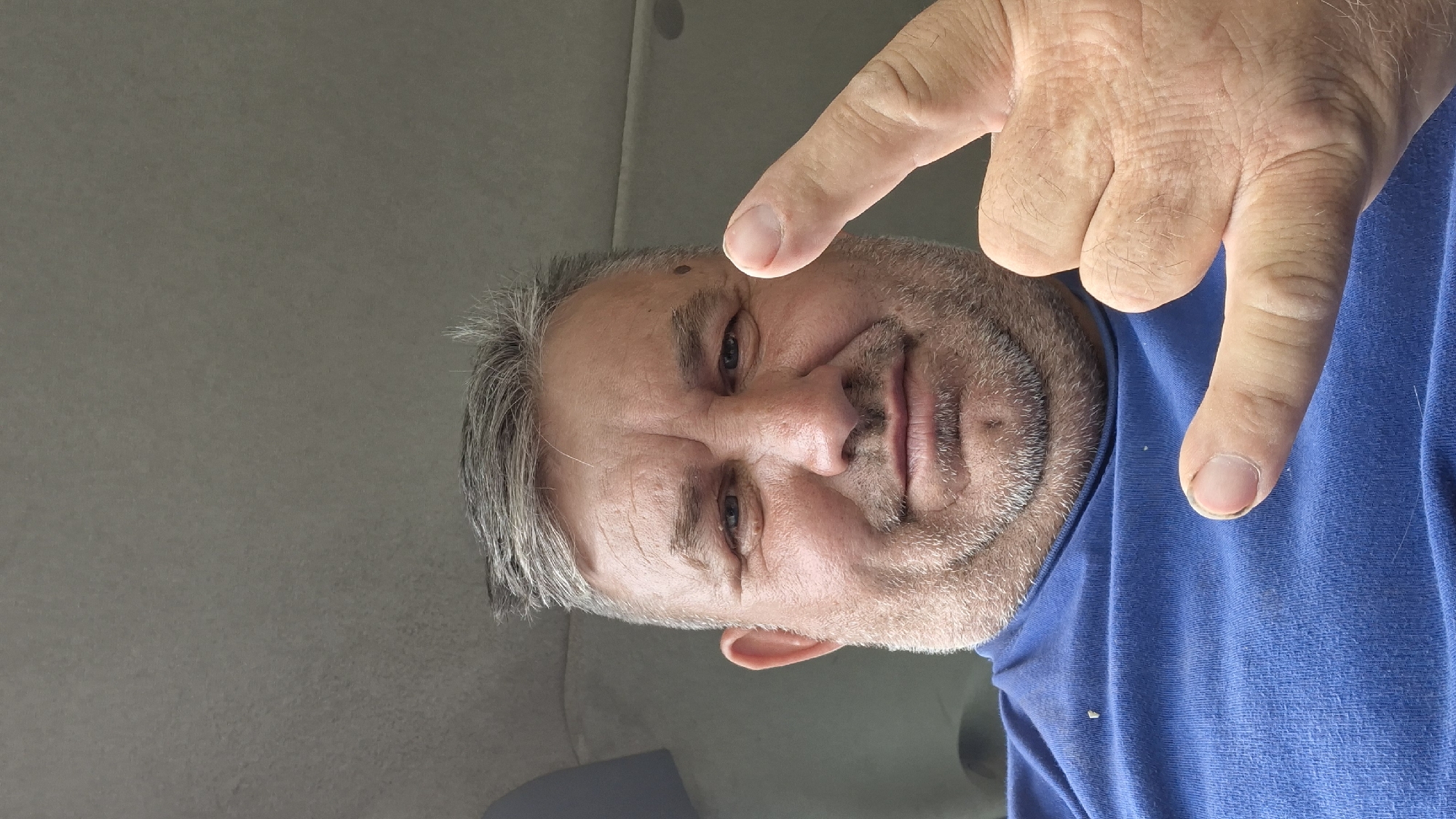 A close-up, selfie-style photograph shows a man with graying hair and a beard. He is wearing a blue t-shirt and has his right hand raised, with his index finger pointing upwards and his thumb and other fingers forming a "shaka" sign. The man has a mole on his left cheek. The background appears to be the interior of a vehicle, with a gray headliner and dark blue paneling visible. The lighting suggests it is daytime. The image is a personal photograph and does not depict any specific location in Hîncești, Moldova.