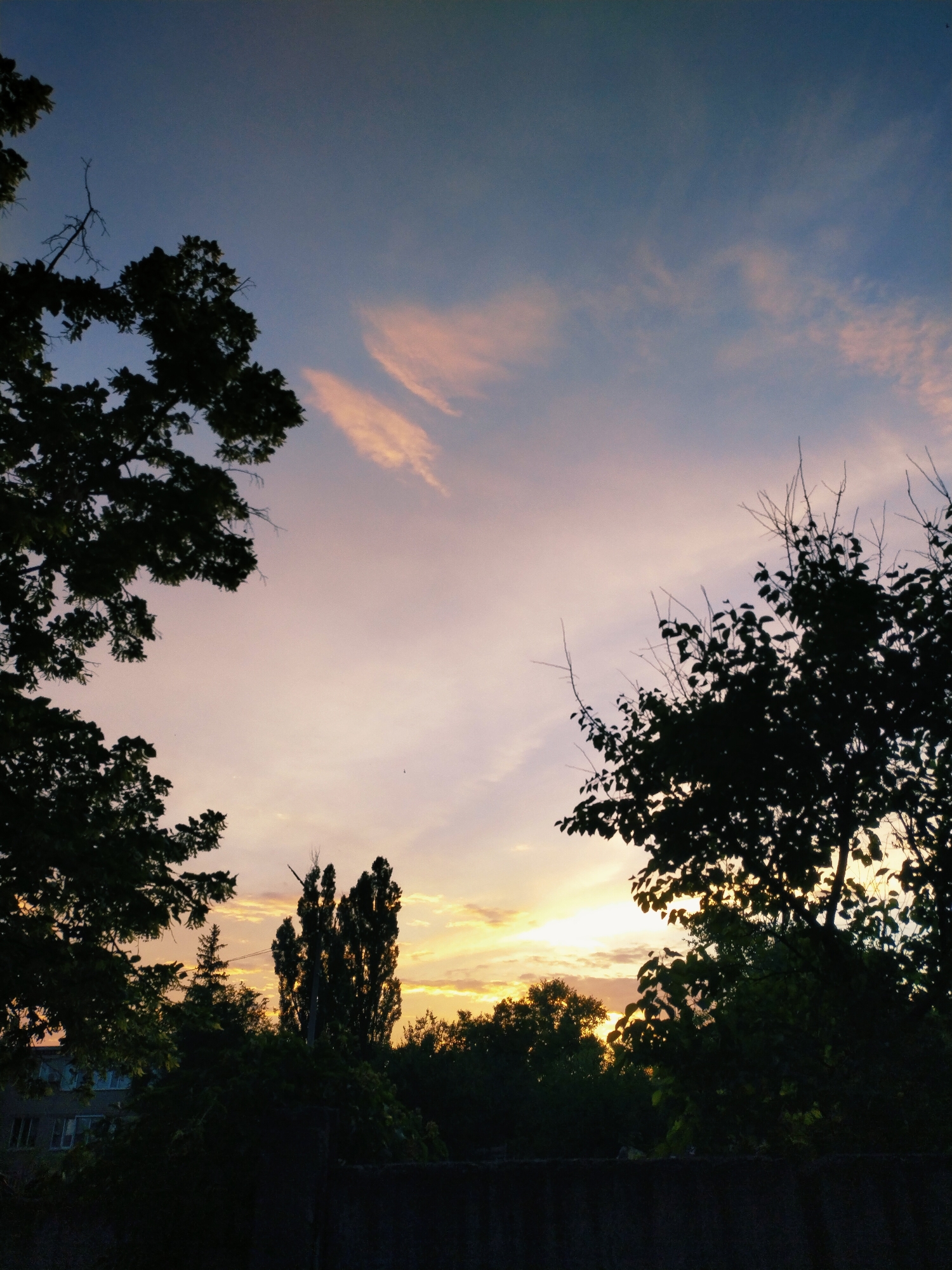 This media file captures a serene sunset scene in Horishni Plavni, Ukraine. The sky is a canvas of soft colors, transitioning from a deep blue at the top to pale pinks and oranges closer to the horizon. Wispy clouds are scattered across the sky, catching the warm glow of the setting sun.
Silhouetted against the vibrant sky are the dark forms of trees and foliage. In the foreground, a dark, textured wall runs horizontally, anchoring the lower part of the image. To the left, the dark leaves and branches of a large tree frame the sky. Further into the scene, a cluster of tall, slender trees stands out, their dark silhouettes stark against the brighter sky. To the far left, a building with visible windows peeks through the greenery, suggesting an urban or suburban edge to this natural setting.
The overall atmosphere is peaceful and tranquil, evoking a sense of calm as day transitions into night. The time of day is clearly sunset, indicated by the specific colors and light in the sky. No people or animals are visible, and there is no visible text in the image. The scene suggests a quiet moment of natural beauty, possibly at the end of a day.