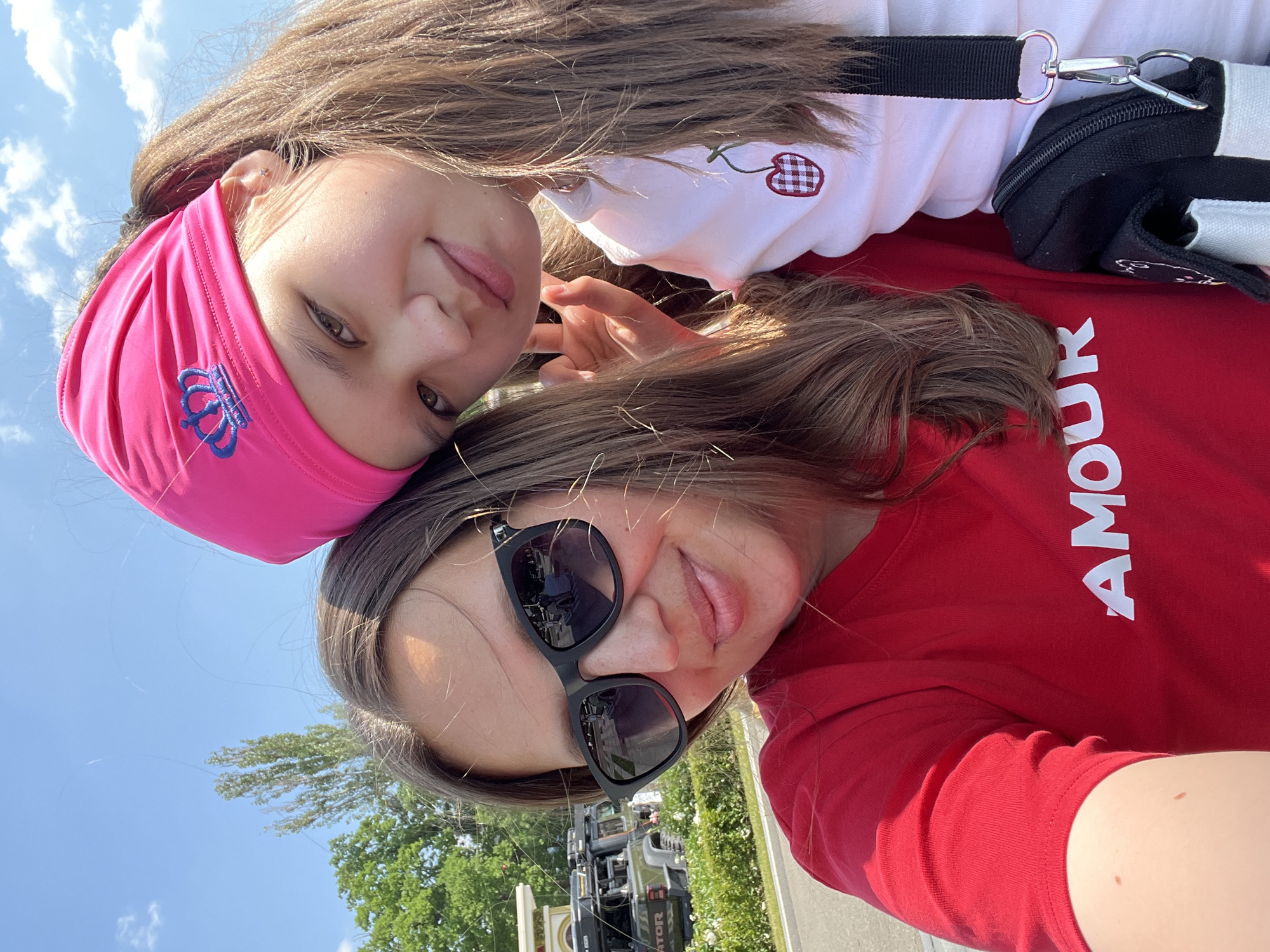 Two females are taking a selfie outdoors. The female on the left, wearing a red t-shirt with "AMOUR" printed on it, has brown hair, sunglasses, and is looking at the camera. The female on the right, wearing a pink headband and a white t-shirt with a cherry design, has brown hair and is also looking at the camera. They are standing in front of a street with trees and vehicles in the background. The environment appears to be a sunny day with a blue sky and some clouds. The location is the city of Moscow, Russia.