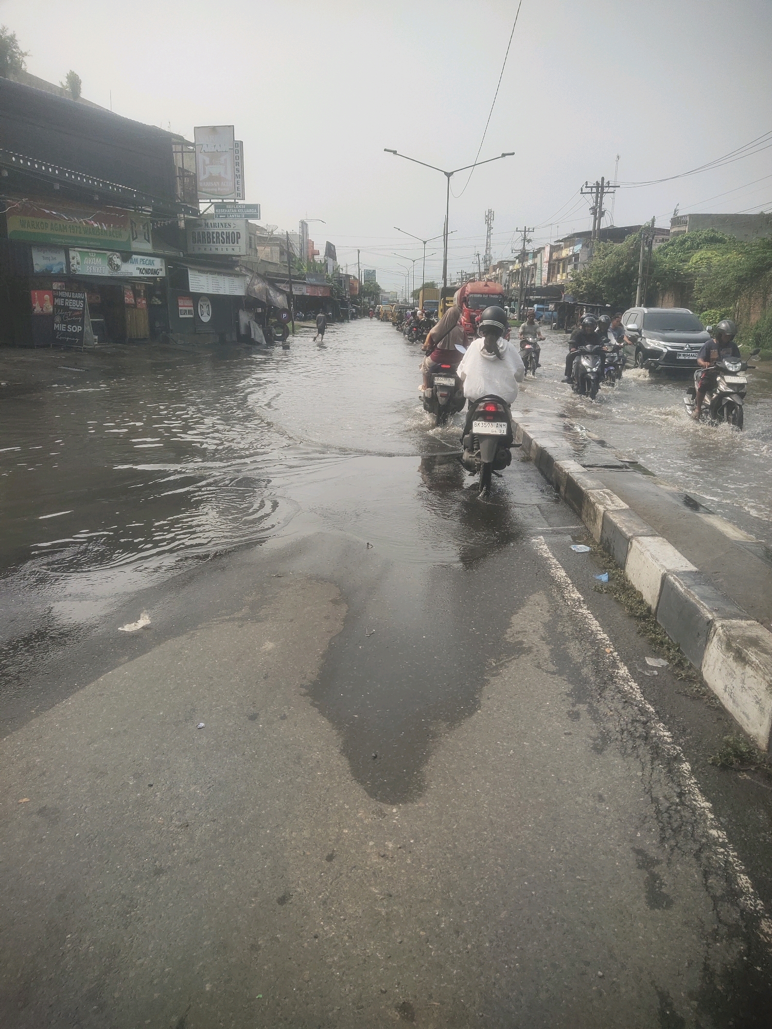 This image depicts a flooded street in the city of Medan, Indonesia, likely during a rainy season. The road is submerged under a significant amount of water, making it difficult for vehicles to pass. Several motorcycles and cars are navigating through the floodwaters, with some riders appearing cautious. On the left side of the street, commercial buildings with various signs are visible, some of which seem to be small businesses or food stalls. The sky is overcast, suggesting continued or recent rainfall. The overall scene conveys a sense of disruption and challenges caused by the flooding in an urban environment.