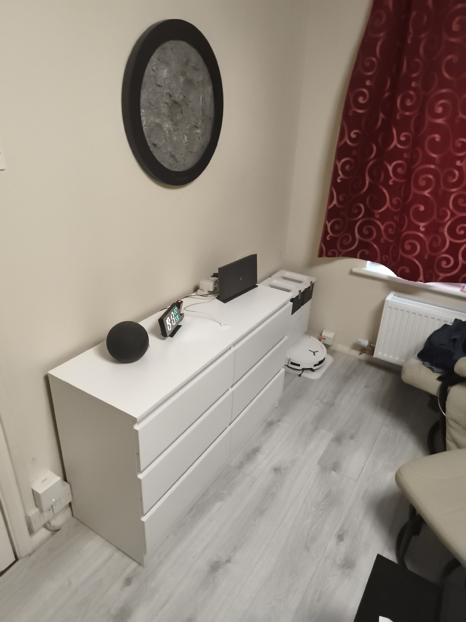 An indoor scene in a room, likely a bedroom or home office, located in Huntingdonshire, United Kingdom. The room features light beige walls and light grey wood-effect laminate flooring.
A long, white six-drawer dresser is positioned against the back wall. On its surface, from left to right, are a black spherical smart speaker, a digital alarm clock displaying "8:00" in green digits, and a closed black laptop. Behind the laptop, a white power strip with various cables is visible. To the left of the dresser, near the floor, a white smart plug is connected to a wall outlet.
Above the dresser, a circular piece of wall art with a black frame depicts a textured, grey, moon-like surface.
To the right side of the image, rich red curtains with a subtle swirling pattern are drawn, partially obscuring a window. Beneath the window is a white radiator. A cream-colored armchair is partly visible next to the radiator. On the floor, near the dresser and radiator, a white Roborock robot vacuum cleaner is docked at its charging and auto-empty station.
The room appears tidy and equipped with modern home electronics, suggesting a functional and lived-in space. No people are present, and no specific activity is taking place. The drawn curtains and artificial lighting imply an indeterminate time of day, possibly evening or early morning, aligning with the "8:00" displayed on the clock.