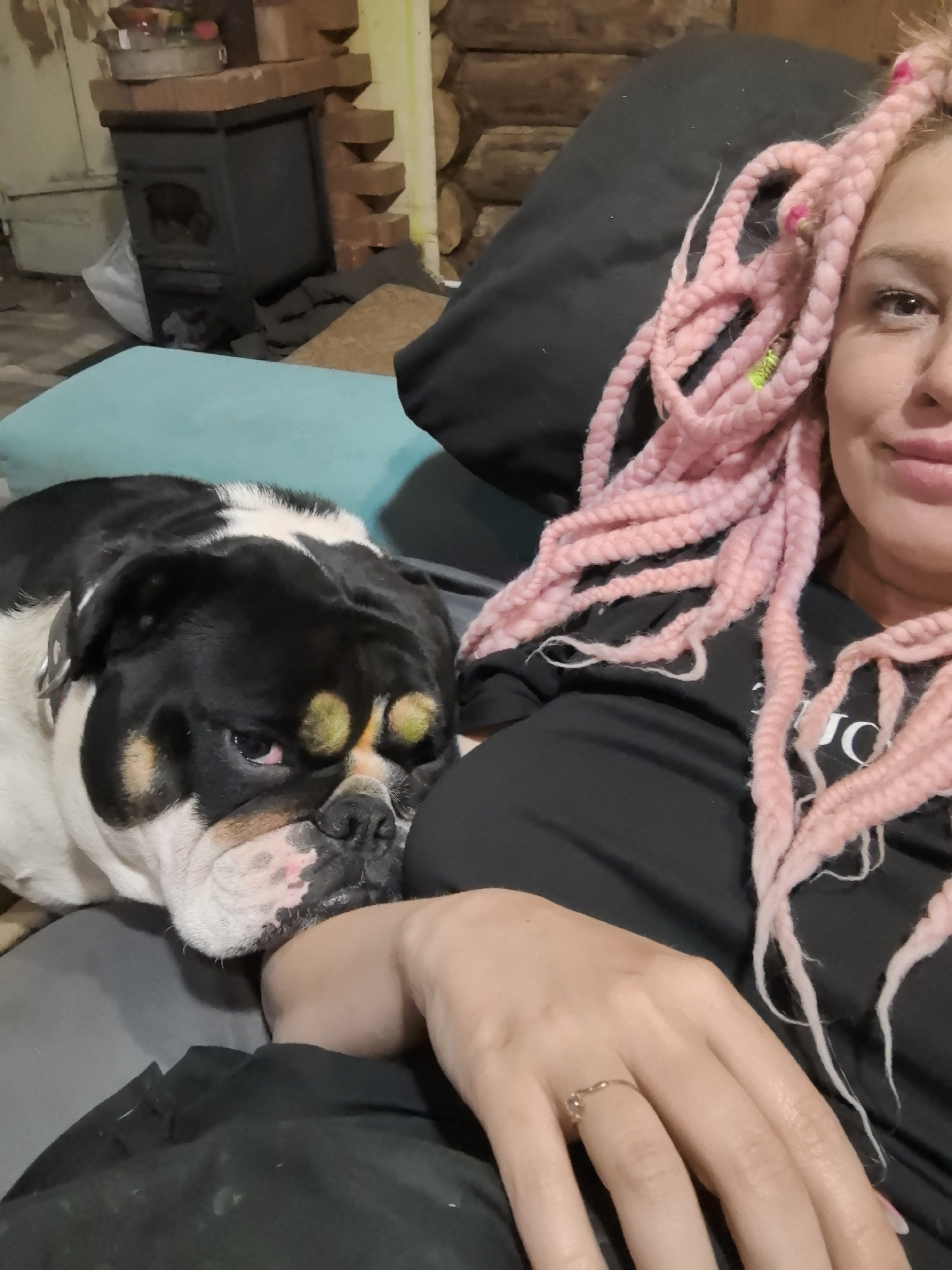 A woman with pink braided hair lies on a bed or couch with a black and white bulldog. The dog rests its head on the woman's arm. The woman is wearing a black shirt. There is a black pillow behind her. A blue pillow is visible behind the dog. In the background, there is a wood-burning stove and a wall of logs. The woman is wearing a ring on her right hand. The location is Noviy, Russia.