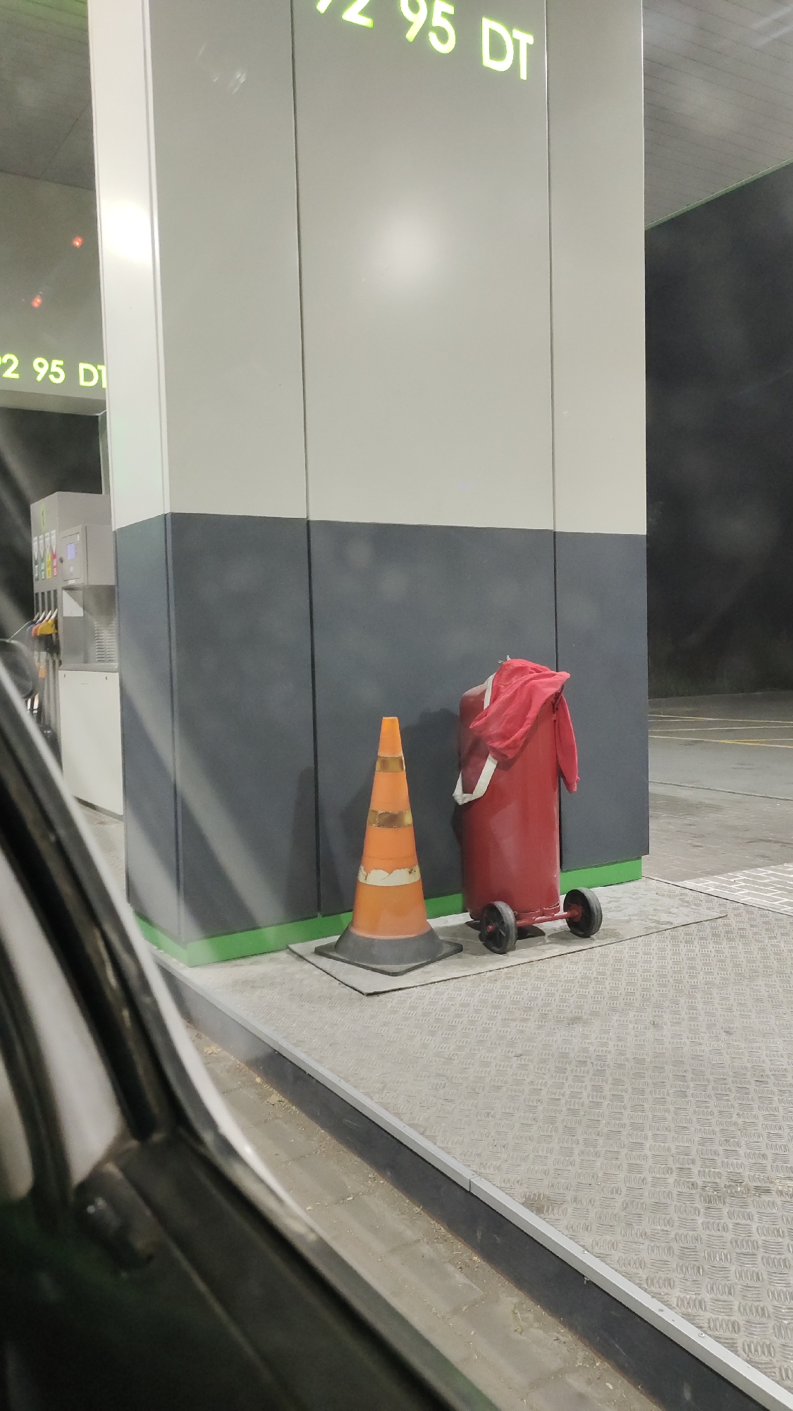 The image depicts a gas station scene at night, with a focus on a red fire extinguisher and an orange traffic cone. The fire extinguisher is cylindrical with two wheels at the base and a red cloth draped over its top. The traffic cone is positioned to the left of the fire extinguisher. In the background, a large grey and white structure, likely part of the gas station canopy, is visible, with illuminated numbers that appear to be prices. The ground is covered with a patterned metal grate. A portion of a car is visible in the foreground, suggesting the photo was taken from inside a vehicle. The environment appears to be outdoors, with a dark sky and a street visible in the distance.