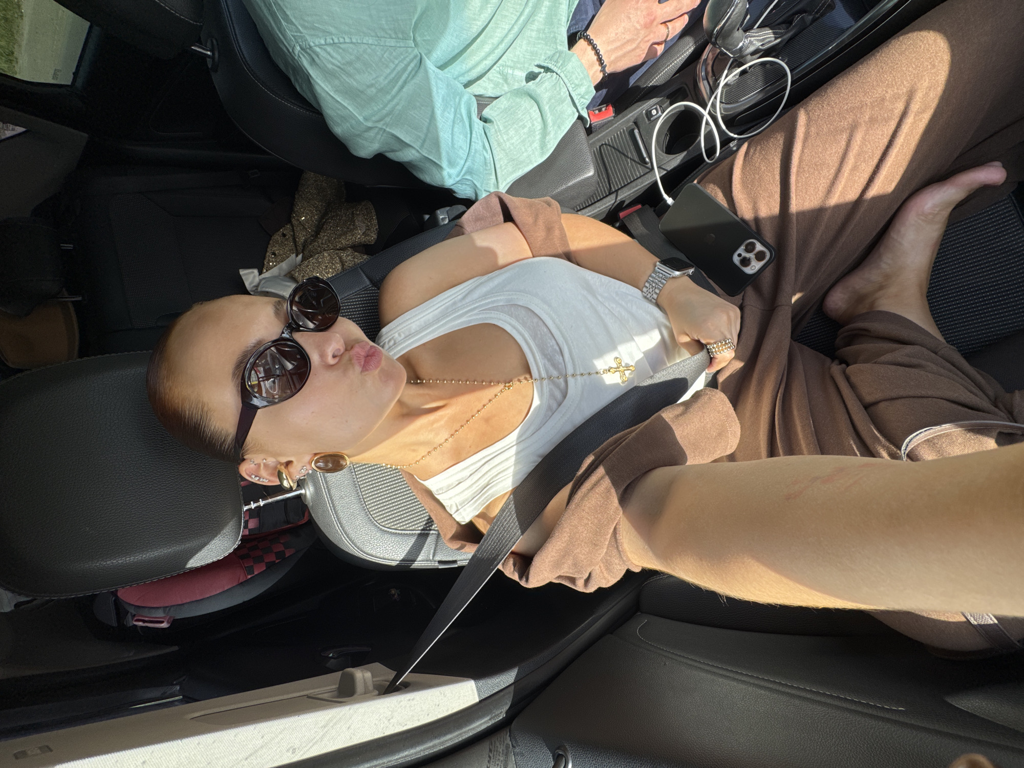 A woman in a car wearing a white tank top, brown pants, gold jewelry, and sunglasses puckers her lips for a selfie. A person in a mint green shirt sits next to her in the passenger seat. A black smartphone and white charging cable are on the center console. The vehicle's seatbelt is fastened. A child's car seat is visible behind the driver's seat. The location is not identifiable as Chaikovsky, Russia.
