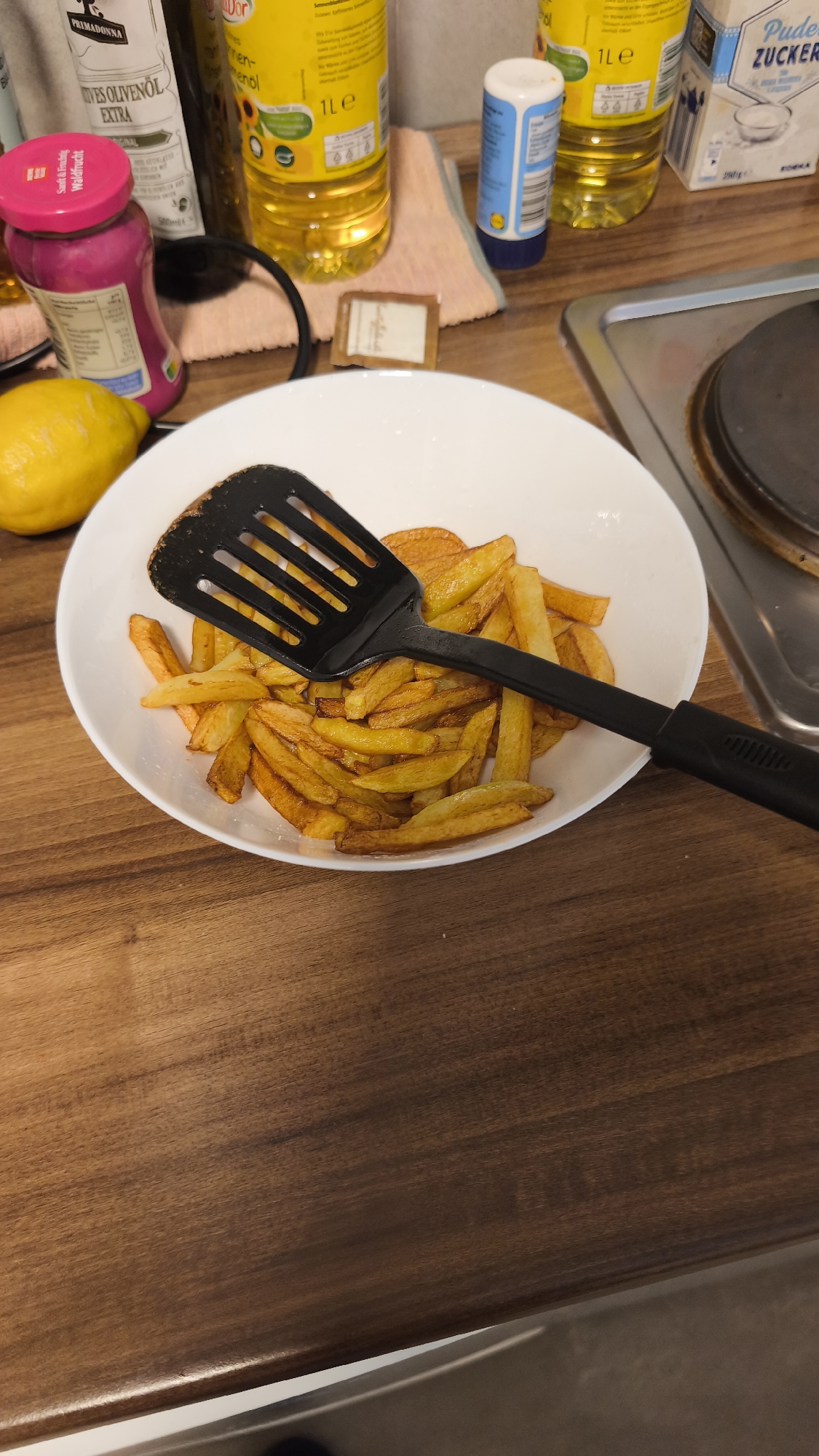 A high-angle, eye-level shot shows a bowl of French fries with a black spatula lying on top. The bowl sits on a wooden countertop, next to a stovetop. In the background, there are several bottles, including bottles of cooking oil and a jar of fruit jam. A lemon is also visible near the bowl. The scene is indoors and appears to be a kitchen setting.