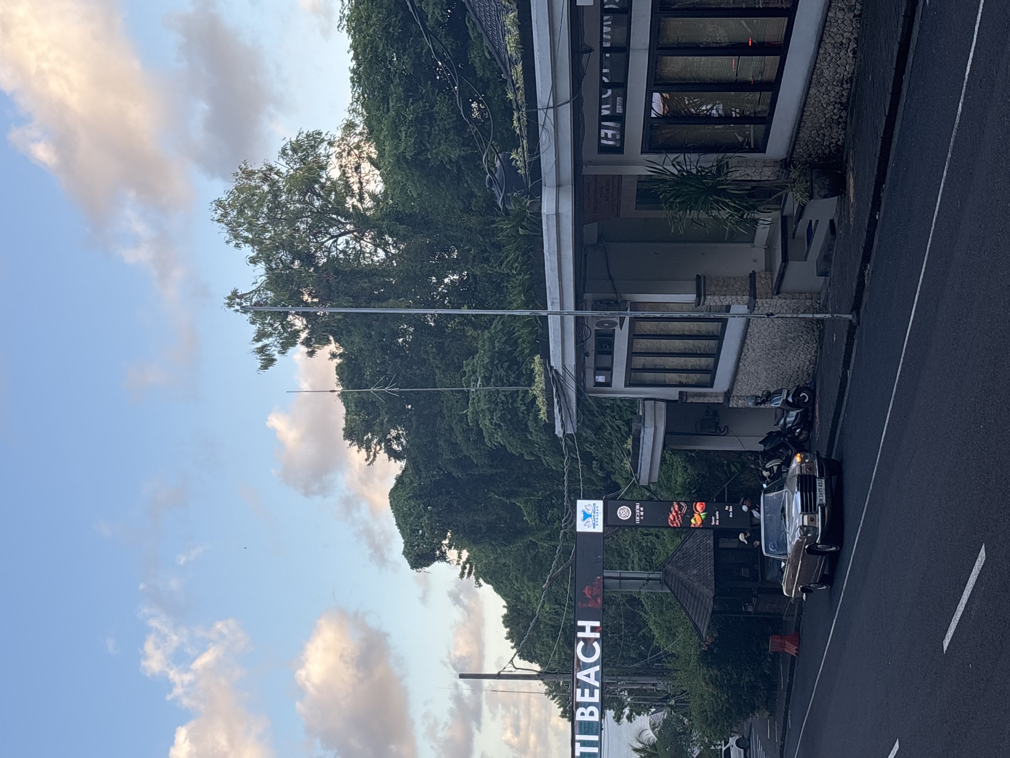 The image shows a street scene in Kuta Selatan, Indonesia. Visible is an old car parked on the side of the road, along with several scooters. A sign reading "Uluwatu Beach" is prominently displayed, indicating the location. A building is on the right with multiple windows and a dark roof. The sky is blue with clouds, and trees surround the area. Several people are partially visible near the buildings.