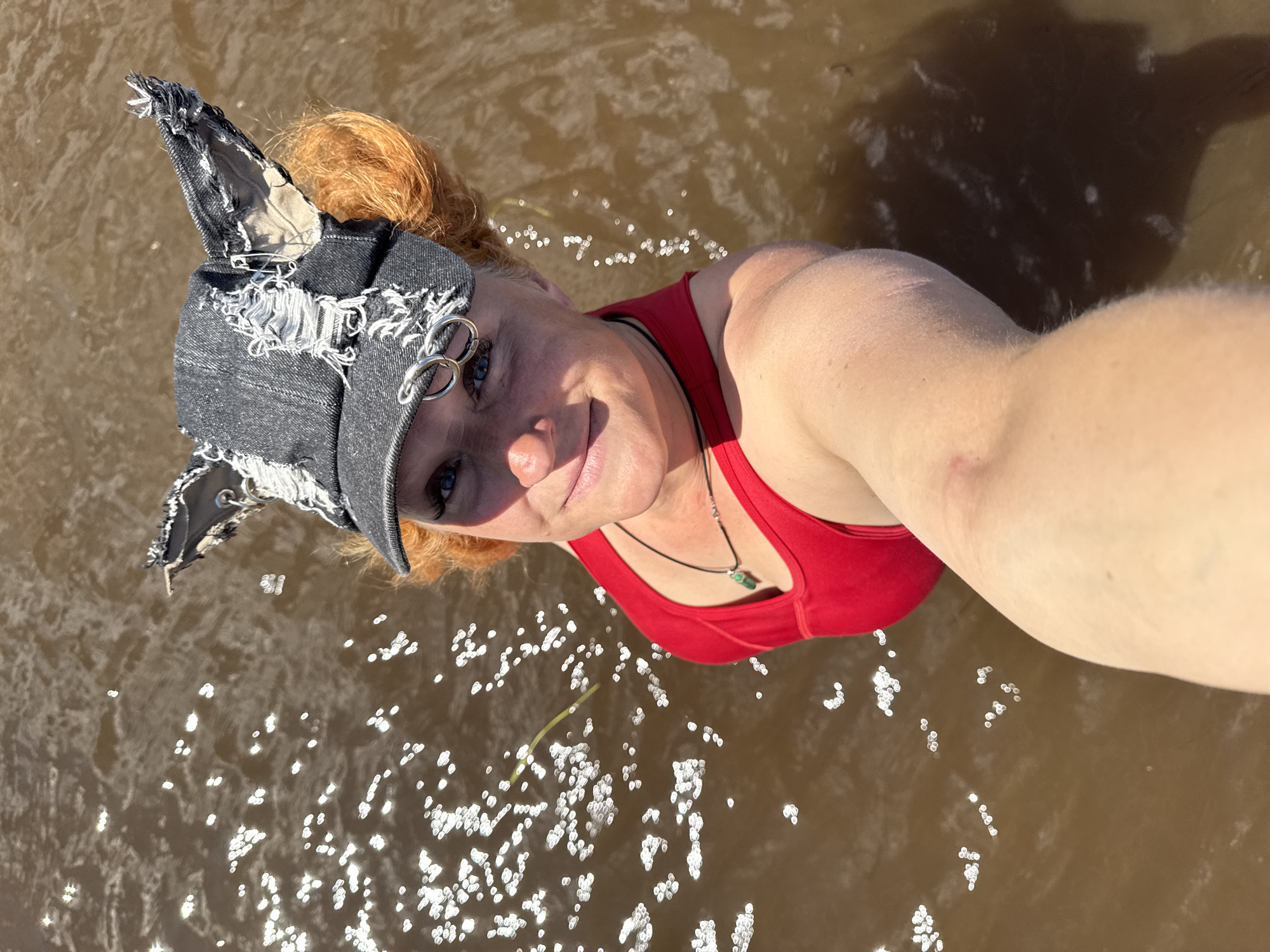 A woman with red hair is taking a selfie in murky, brown water. She is wearing a red tank top and a distressed denim hat with holes and frayed edges. The hat is decorated with safety pins and large metal rings. She is also wearing a necklace with a green pendant. The water is reflecting sunlight, creating bright, sparkling highlights. The woman appears to be smiling. The setting suggests an outdoor activity, possibly in a river or lake. The image is taken from a high angle, looking down at the woman. The lighting suggests it is daytime, likely sunny. There is no visible text in the image.