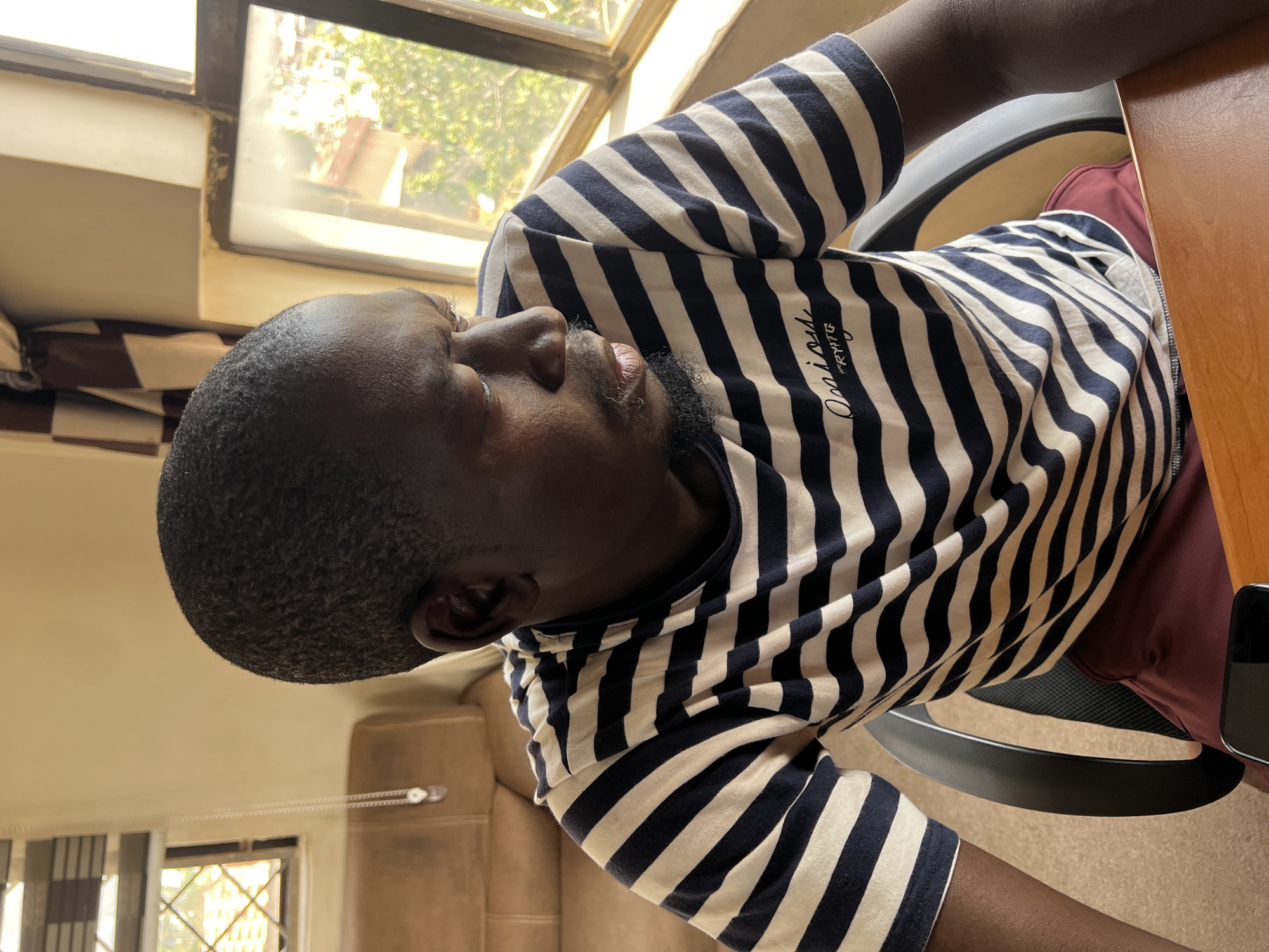 In Jos, Nigeria, a man with dark skin and short hair is seated in a chair, looking to his right. He is wearing a horizontally striped t-shirt with navy blue and cream bands, and a small embroidered logo on the left side of his chest reads "Enjoy" above "RYTG". His lower body is clad in maroon shorts. The man is positioned in front of a wooden desk, on which a dark-colored smartphone is visible. Behind him, to his left, a tan leather sofa is present. The environment is illuminated by natural light filtering through a window with a diamond-patterned grate. A patterned curtain, featuring alternating dark brown and white stripes, hangs to the left of the window. The background beyond the window shows buildings and greenery, suggesting an outdoor urban setting.