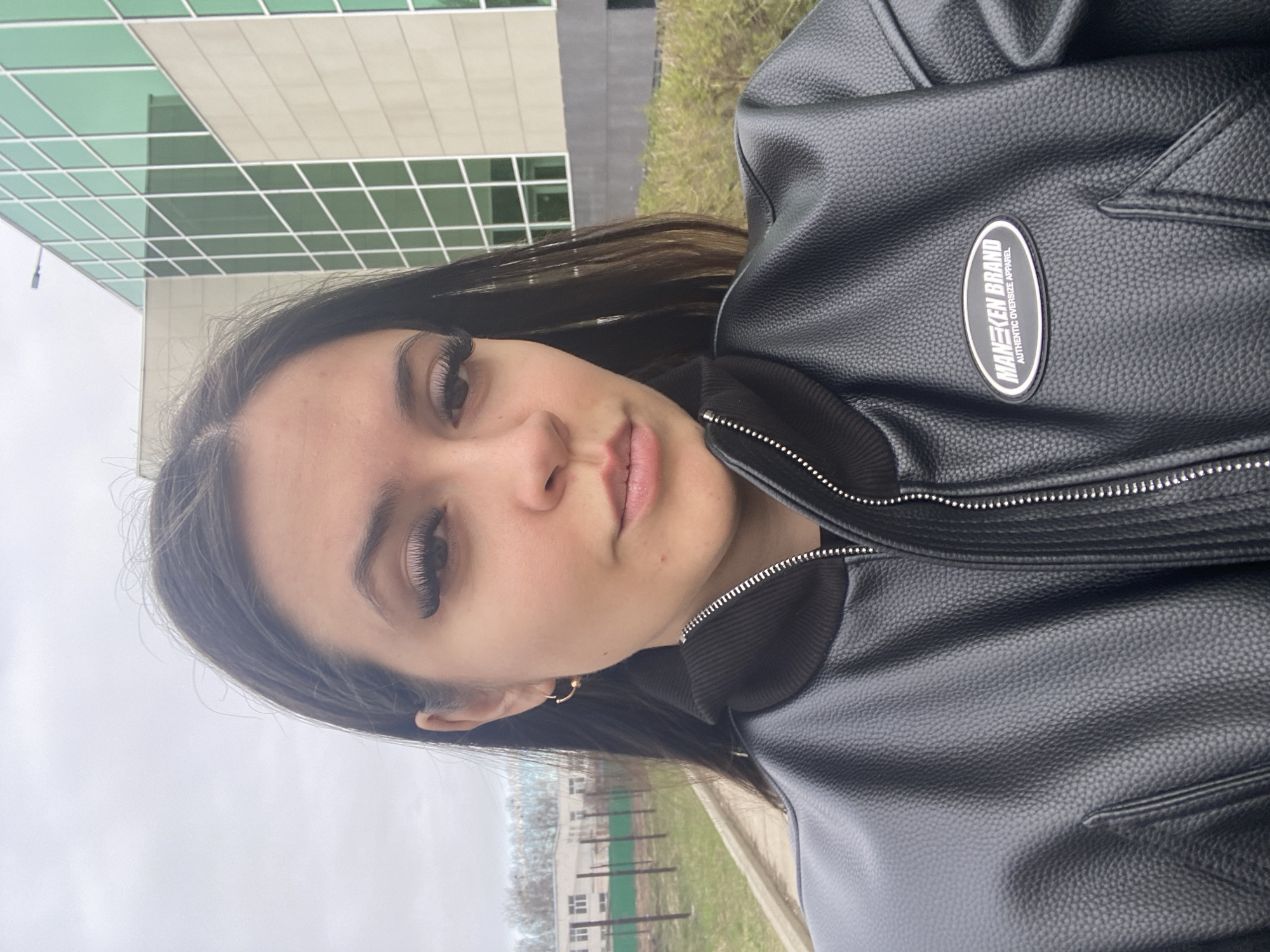 A woman is facing the camera, wearing a black leather jacket with a “MANKEN BRAND” logo on the right chest. She has dark hair and eyelashes, and is wearing small gold hoop earrings. The background consists of a modern building with large windows and green grass. The location appears to be an outdoor area, possibly in the city of Chaikovsky, Russia, based on visual cues of architecture and vegetation.