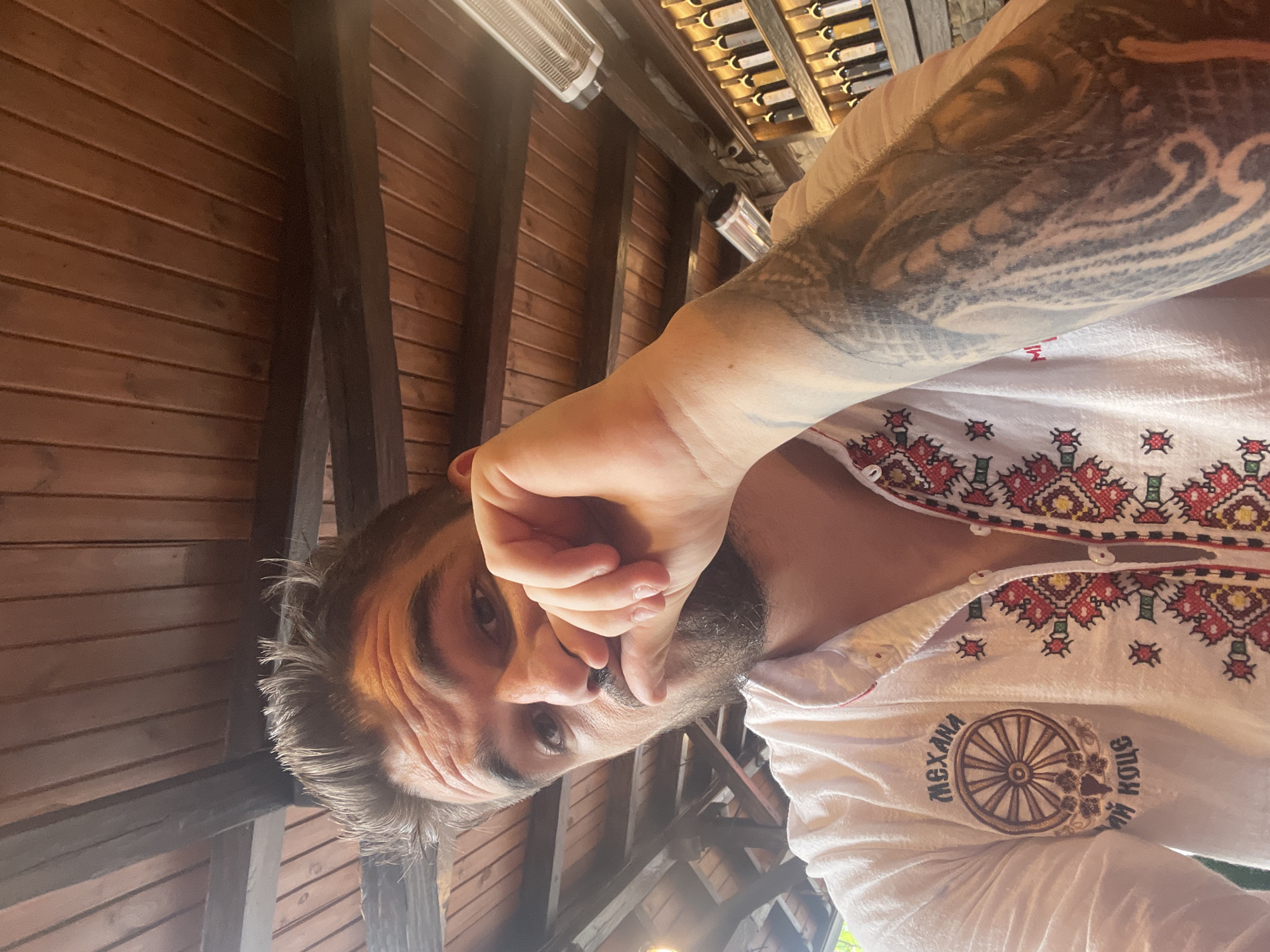 A man is depicted in a portrait-style shot, rotated 90 degrees clockwise. His face is positioned towards the bottom of the frame, with his body extending upwards. He has a dark beard and dark hair, and his right hand covers his mouth and chin. His eyes are looking directly towards the viewer. His left arm, prominent in the foreground, is heavily tattooed with intricate dark designs. He wears a white shirt adorned with traditional red, black, and gold folk-style embroidery across the chest and shoulders. On the left side of the shirt, a circular emblem resembling a wooden wagon wheel is visible, surrounded by Cyrillic text that reads "МЕХАНА" (Mehana) and "ХОВ" (Hov).
The background suggests a rustic indoor or covered outdoor setting. It features a wooden plank ceiling or wall with dark support beams running across the frame (if viewed upright). A silver radiant heater is mounted on one of the beams on the left side. In the upper right corner, a wooden rack holds several bottles horizontally, likely wine. The lighting is somewhat dim, implying an indoor environment or shade during the daytime. The man's pose, with his hand partially obscuring his face, creates a thoughtful or casual mood.