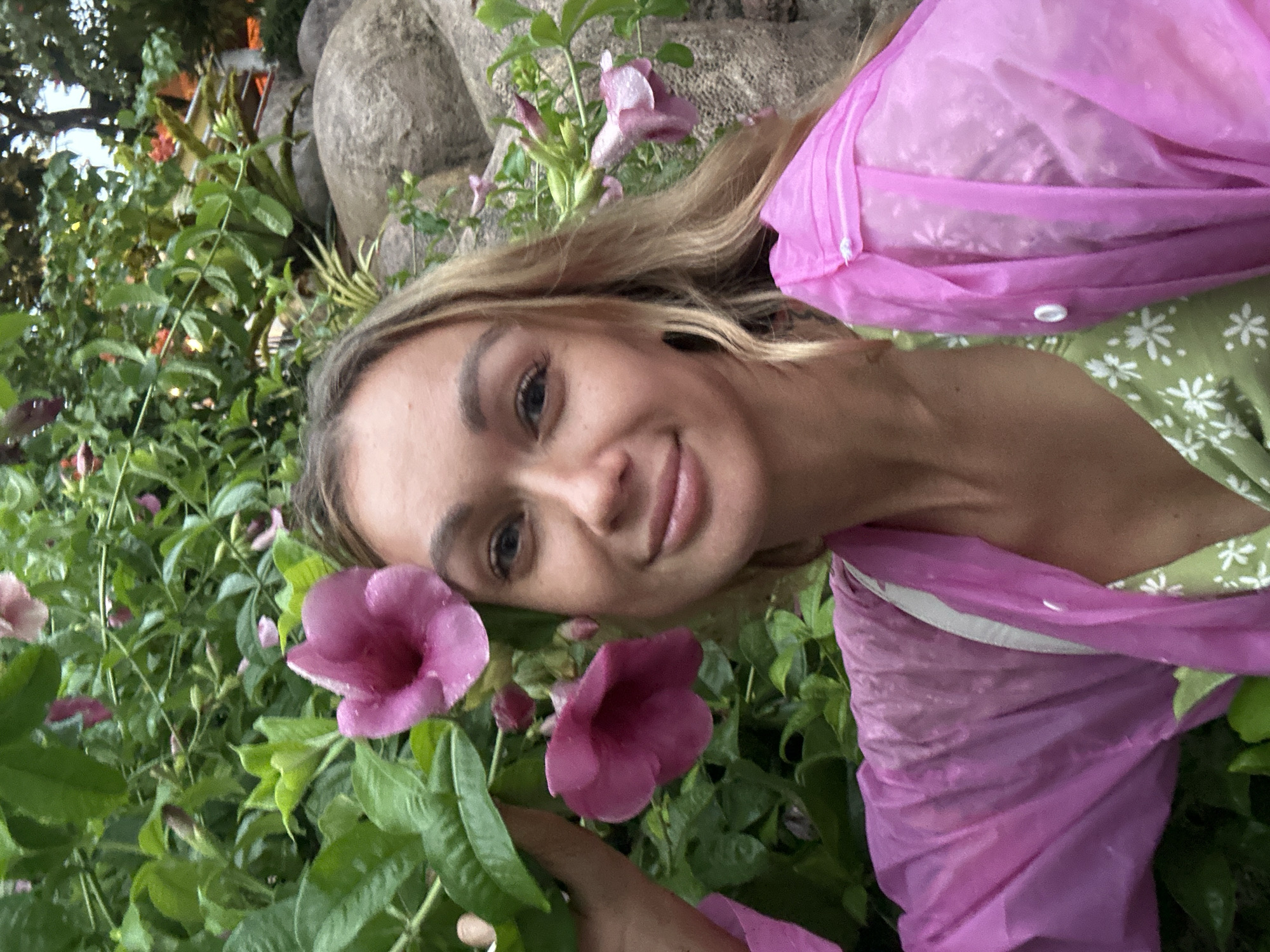 A woman poses with pink flowers in a garden, wearing a pink poncho over a green dress. The location appears to be in a park setting in Phu Thuy Ward, Vietnam, as suggested by the background flora and foliage.