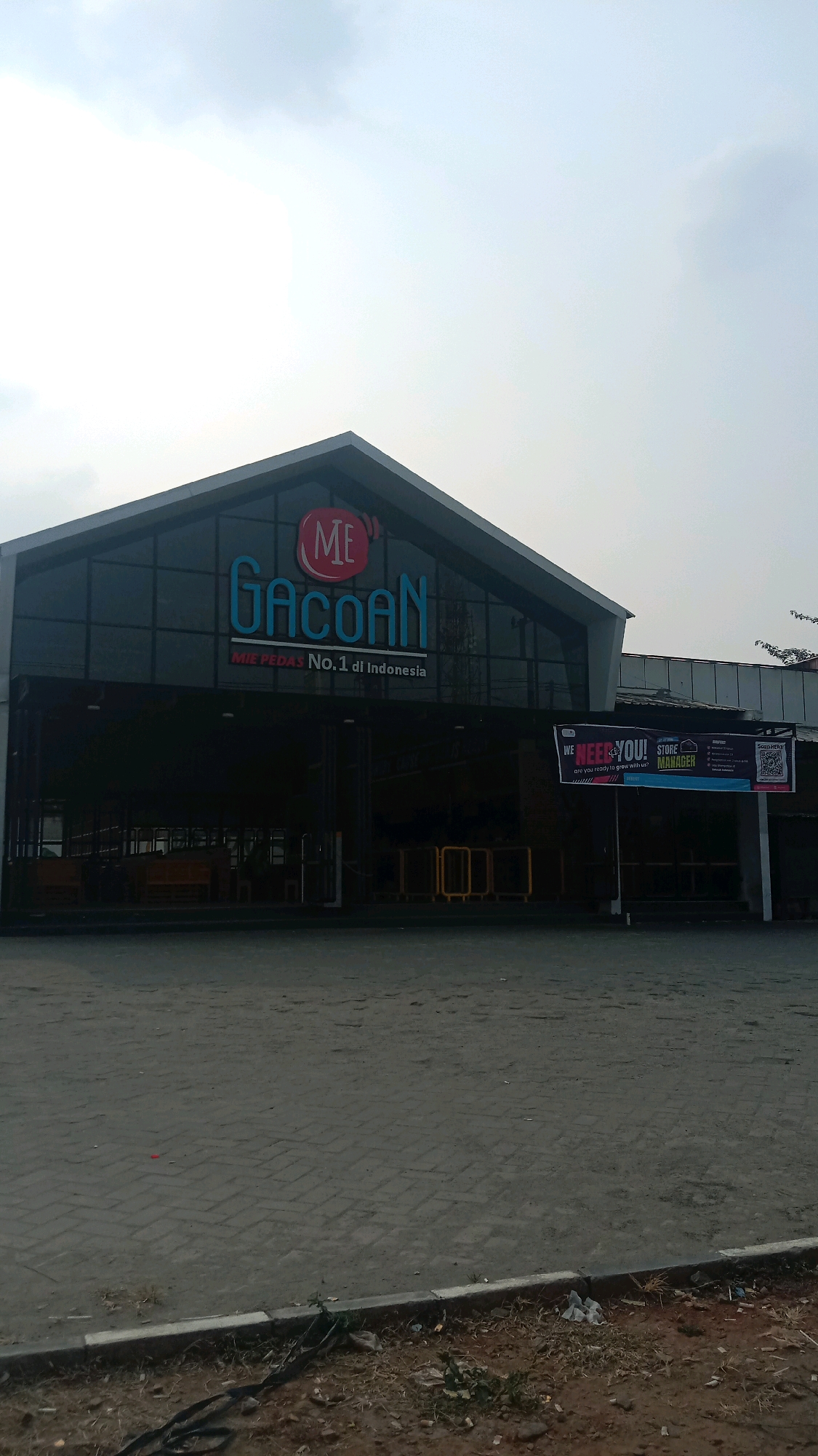 The image shows the exterior of a restaurant with a prominent sign reading "ME Gacoan". Below this, smaller text states "MIE PEDAS No.1 di Indonesia". The building has a modern, angular design with large glass windows. To the right of the entrance, a banner advertises for a "STORE MANAGER" position, stating "WE NEED YOU! are you ready to grow with us?". The foreground consists of a paved area with some dirt and debris. The sky is overcast. The context provided indicates this location is in the city of Bekasi, Indonesia.