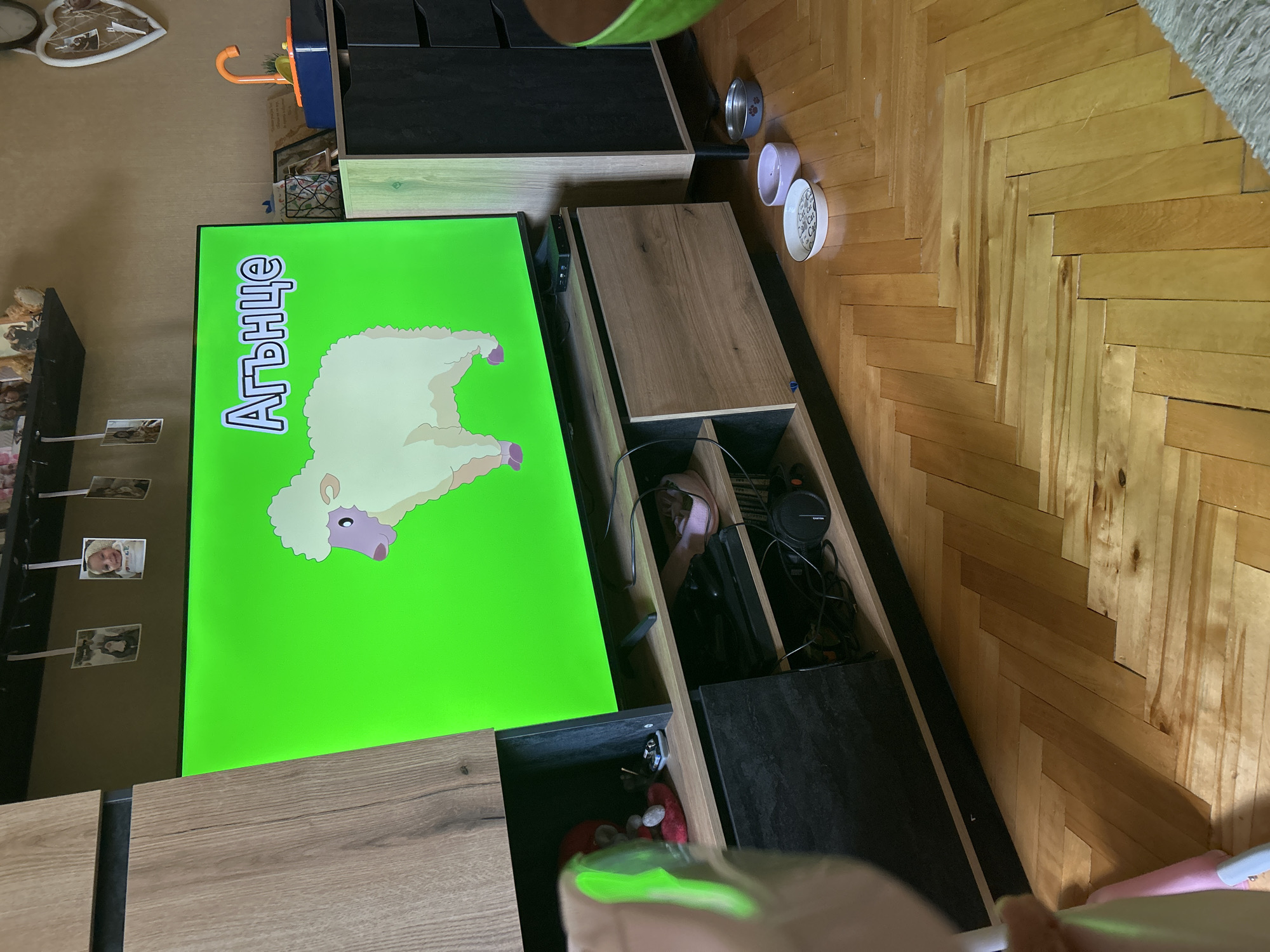 The image depicts a living room scene, likely in Sofia, Bulgaria, centered around a large television screen displaying a cartoon sheep with the Bulgarian word "Агънце" (lamb) above it. The TV is positioned on a wooden entertainment unit, which is part of a larger furniture arrangement. The room features a herringbone-patterned wooden floor. On the floor, near the entertainment unit, are three pet bowls, suggesting the presence of pets. The background shows a wall with some decorative items and a shelf above the TV with several small framed photos. To the right of the TV, a portion of another piece of furniture, possibly a dresser or cabinet, is visible, with a toy or electronic device on top. The overall impression is of a comfortable, lived-in domestic space.
