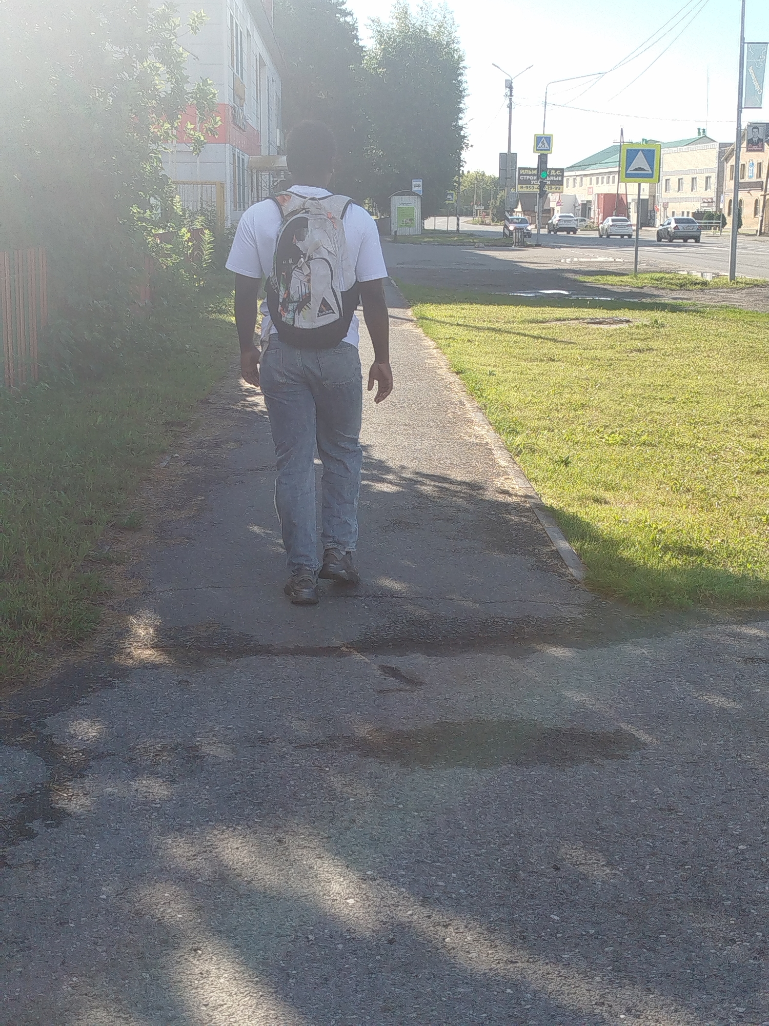 A rear view of a Black man wearing a white t-shirt and light blue jeans walks down a sidewalk on a sunny day. He is carrying a gray backpack with a colorful graphic design on it. The sidewalk is made of asphalt and is partially covered in shadows. To the left of the sidewalk is a grassy area, and to the right is a street with several cars driving. In the background, there are buildings and trees. There are also traffic lights and street signs visible.