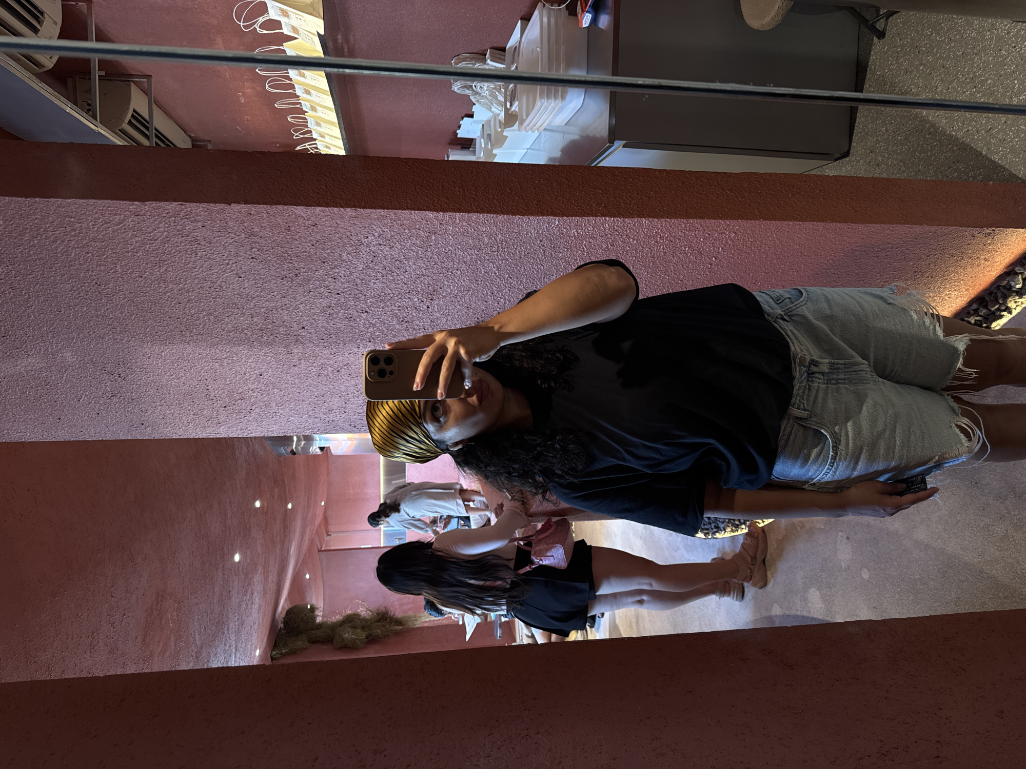 A person is taking a selfie in a mirror, with the reflection showing an interior space with a pink-toned wall. A second person is reflected behind the selfie-taker, wearing a black skirt and carrying a pink purse. Other people are visible in the background. The mirror reflects the restaurant's ceiling and the air conditioning system. The location is North Kuta, Indonesia.