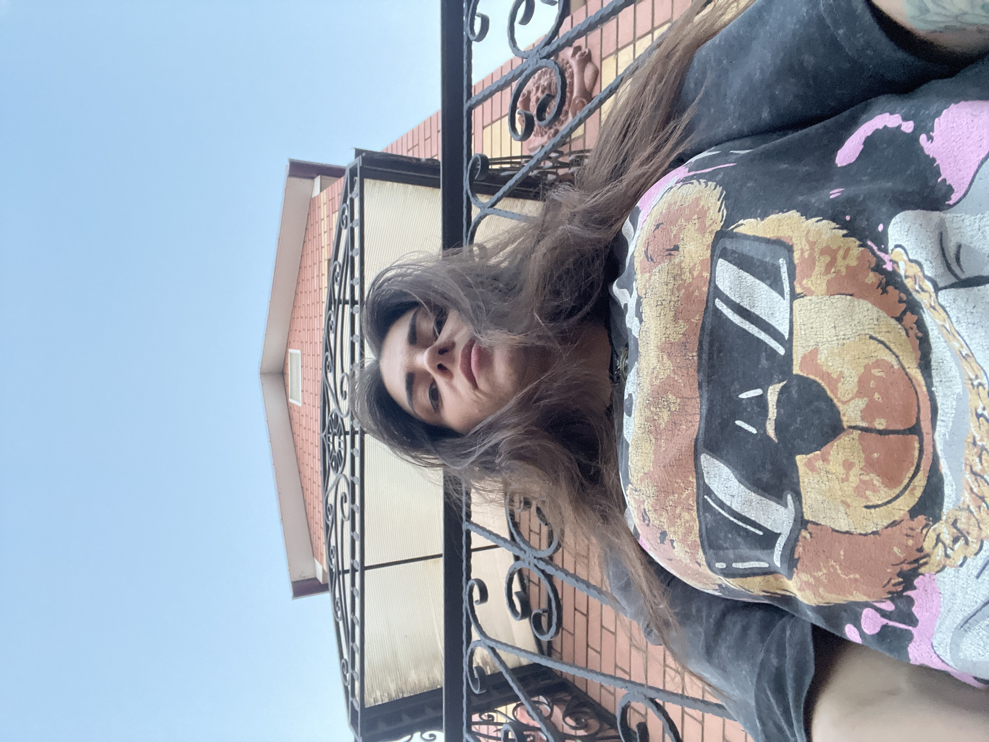 A low-angle, outdoor, selfie-style photograph shows a woman with long, dark hair, wearing a dark t-shirt with a graphic of a teddy bear wearing sunglasses. She is standing on a balcony or porch with a black wrought-iron railing in the foreground. Behind her, a red brick building with a white awning and a red-tiled roof is visible. The sky above is clear and blue. The location appears to be a residential area.