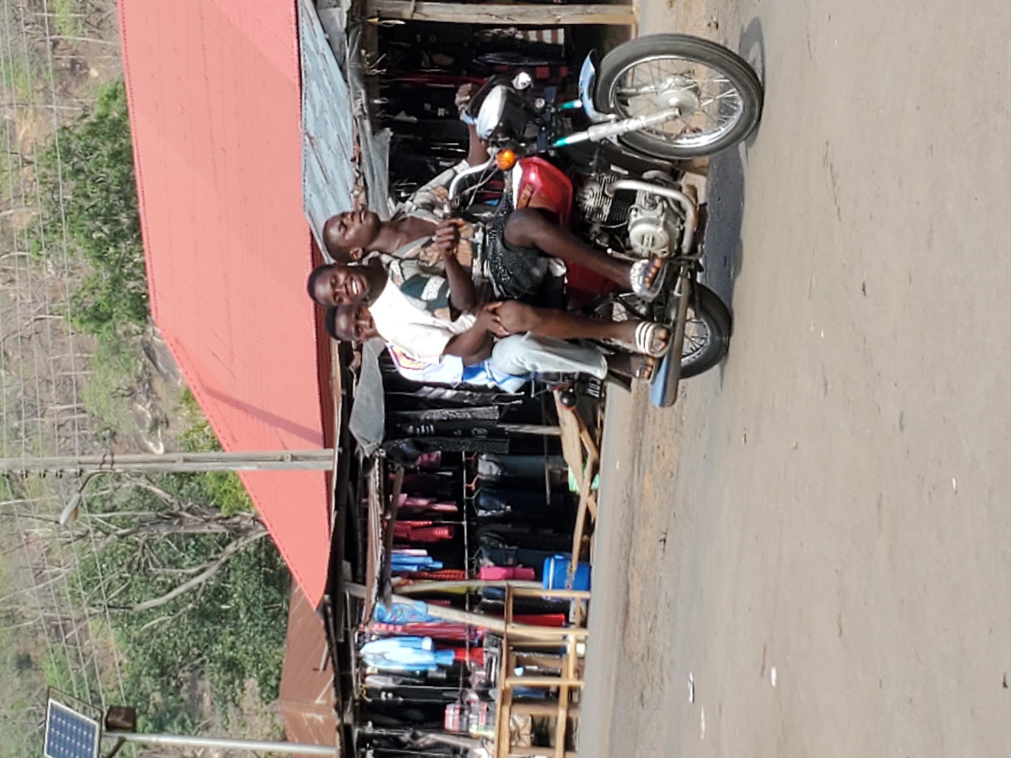 The media file captures three young men seated on a red motorcycle on a sunny day in Mararaba, Nigeria. The motorcycle is stationary on a dirt or unpaved road. All three individuals are smiling or laughing broadly, looking towards the camera, conveying a cheerful mood. The man in the rider's position wears a patterned shirt and dark shorts. The middle passenger is dressed in a white t-shirt over a light blue long-sleeved shirt and light-colored trousers. The third passenger, at the back, wears a dark t-shirt.
To the left of the motorcycle, an open-fronted market stall with a bright red corrugated metal roof is visible. The stall displays various items such as bags, plastic containers, and other goods, indicating a local market setting. In the foreground on the far left, a small solar panel is mounted on a pole, likely for street lighting, next to a simple light fixture. The background features green trees and distant hills, suggesting a semi-urban or rural landscape. The bright sunlight casts strong shadows, consistent with a clear day. No discernible text is visible in the image.