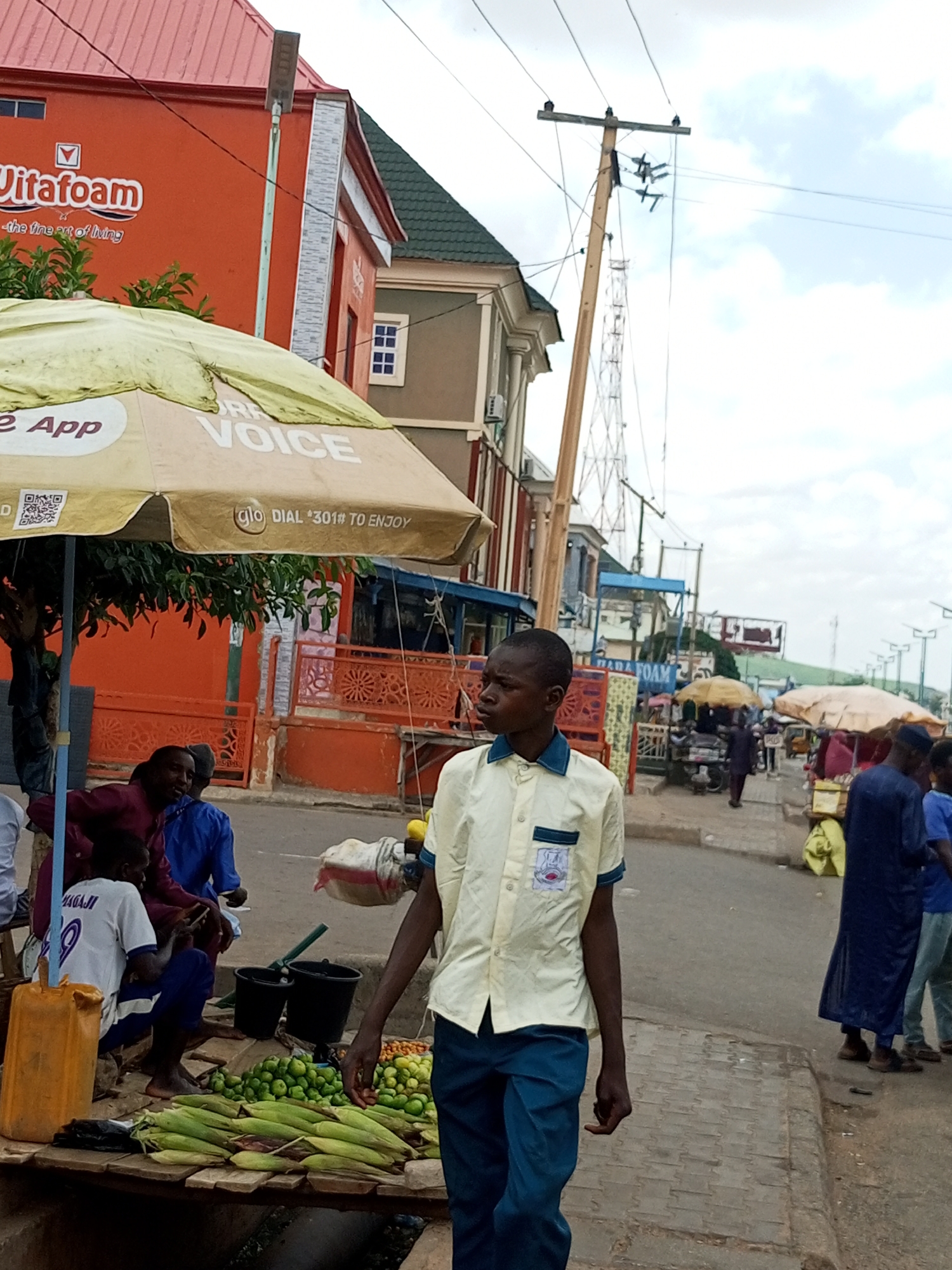 The image depicts a street scene in Arawa, Nigeria. In the foreground, a young boy wearing a light yellow collared shirt with a blue trim and a blue emblem on the pocket, and dark blue trousers, stands facing to the left, looking away from the camera. His right arm is extended slightly forward and his hand is open. To his left, a wooden stall displays an abundance of fresh produce, including corn on the cob, small green fruits, and what appear to be tomatoes. Two black buckets are positioned behind the produce. A bright orange plastic container is also visible near the stall.
Behind the stall, several people are gathered. One man in a maroon shirt and another in a blue shirt are interacting. A boy in a white t-shirt with the number "20" and the name "MAGAJI" is seated, looking at a smartphone.
The background reveals a bustling market or street with several stalls, some covered by umbrellas. Buildings with orange and green roofs line the street. A large orange building with the text "Vitofoam" is prominent on the left. Electrical poles with wires crisscross the sky. The overall environment suggests a lively commercial area in a Nigerian city.