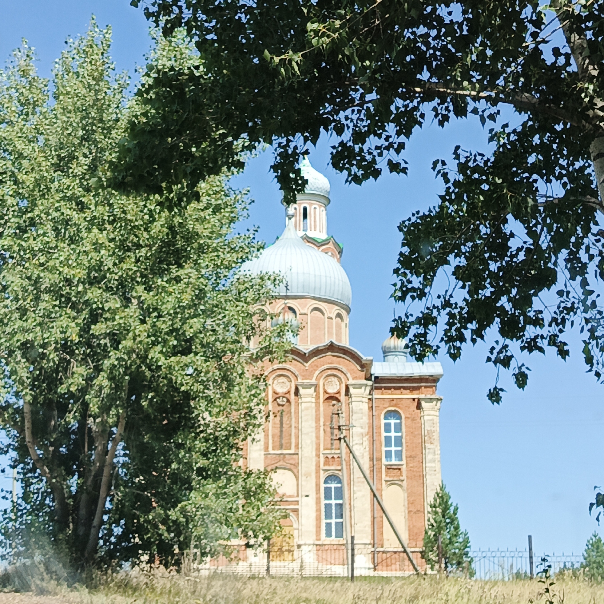 A brick church with a light blue dome is partially obscured by the branches of trees. The church has arched windows with white mullions and decorative carvings. The sky is clear and blue. The church appears to be located in a rural area, with dry grass and a metal fence visible in the foreground. The image is an exterior shot on a sunny day.