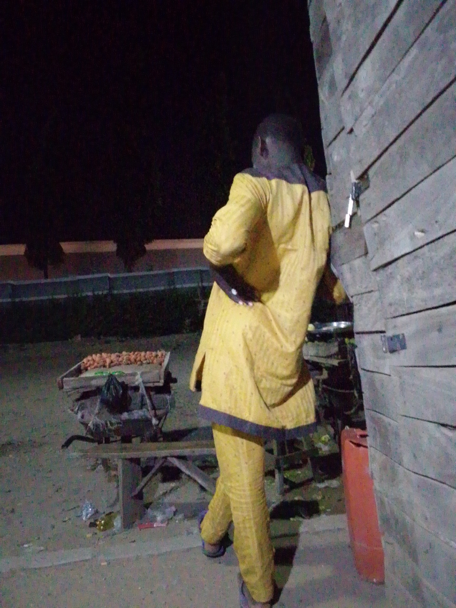 In Ajaokuta, Nigeria, at night, a person wearing a yellow outfit stands near a wooden structure, next to a cart loaded with objects, possibly produce. Debris litters the ground.