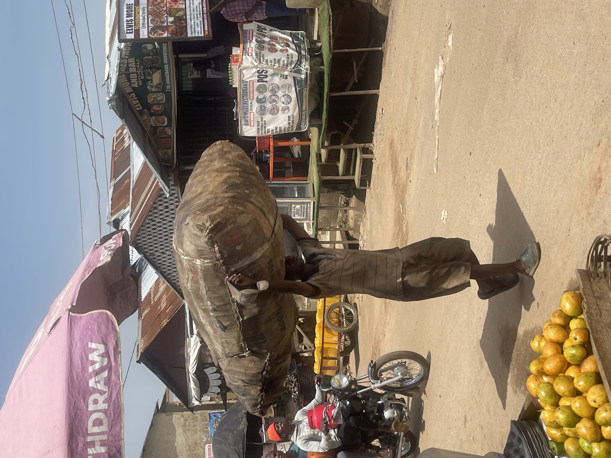 The image depicts a street scene in Abuja, Nigeria. A man is carrying a large, elongated, brown object on his shoulder, possibly a bundle wrapped in cloth or a large container. He is wearing dark trousers and a light-colored shirt. In the background, there are street vendors, one selling a pile of yellow and green fruits, possibly mangoes. A motorcycle is parked nearby. There are several storefronts and signs visible, including one for "ELVIS MORE RESTAURANT," with the restaurant name and menu items listed. A pink awning is partially visible on the left. Overhead, power lines run across the sky. The overall scene appears to be a bustling commercial area.